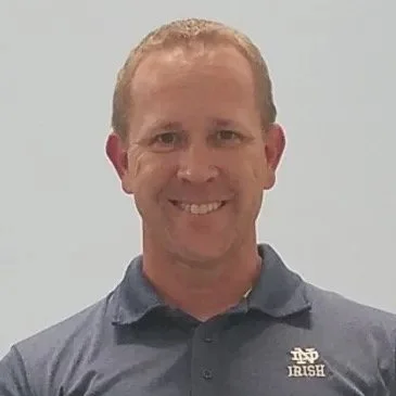 A man with short light brown hair smiling, wearing a dark gray polo shirt with a Notre Dame logo and the word 'Irish' on it, standing against a plain light gray background.