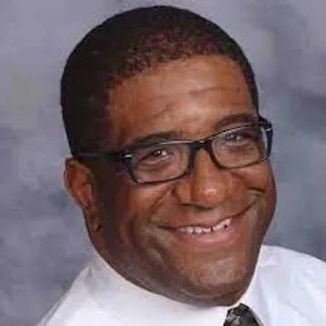 A smiling man with dark skin, glasses, and short black hair wearing a white shirt and a tie, against a gray background.