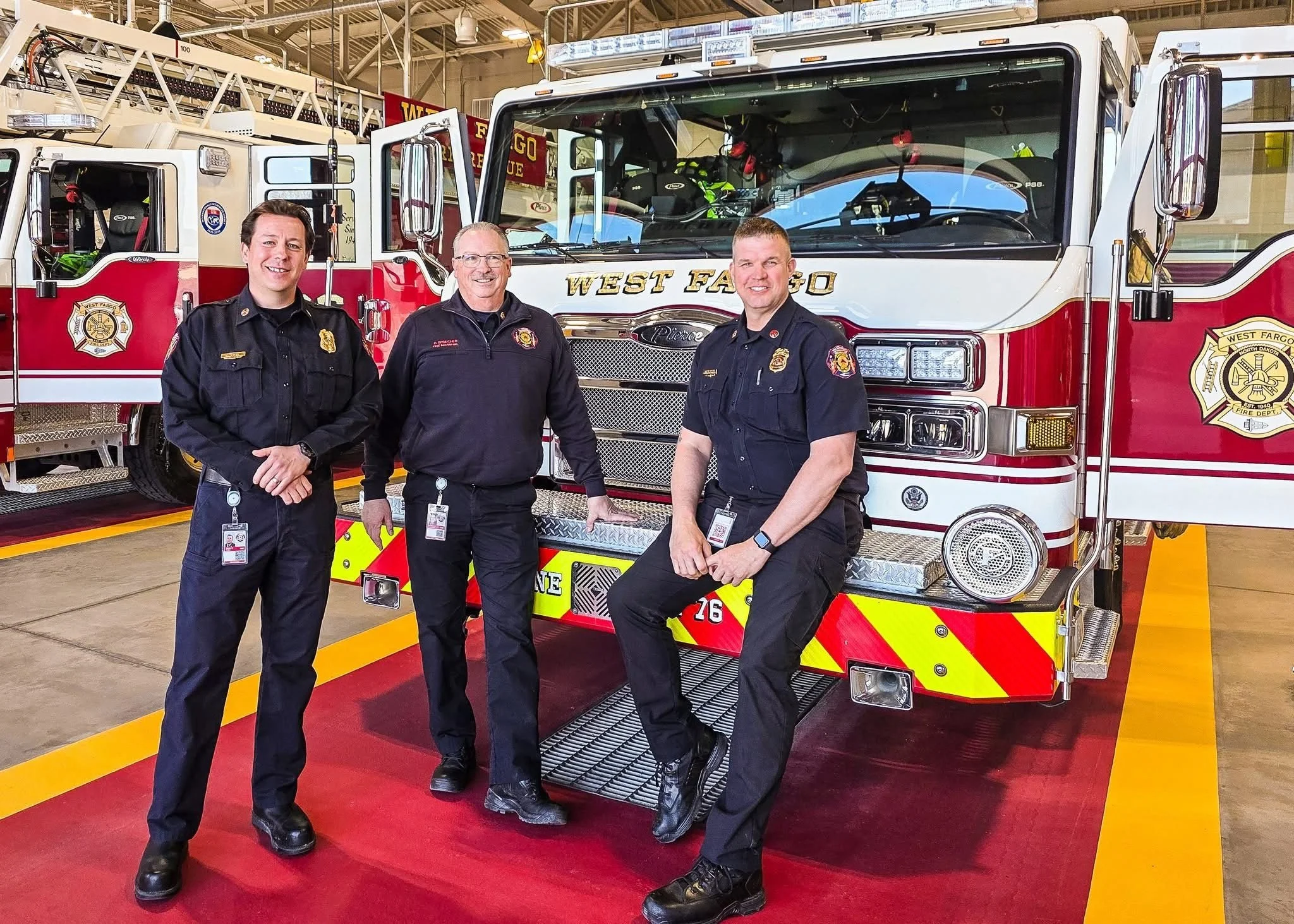 Three firefighters stand inside a fire station next to a red and white fire truck. They are dressed in black uniforms and smiling at the camera.
