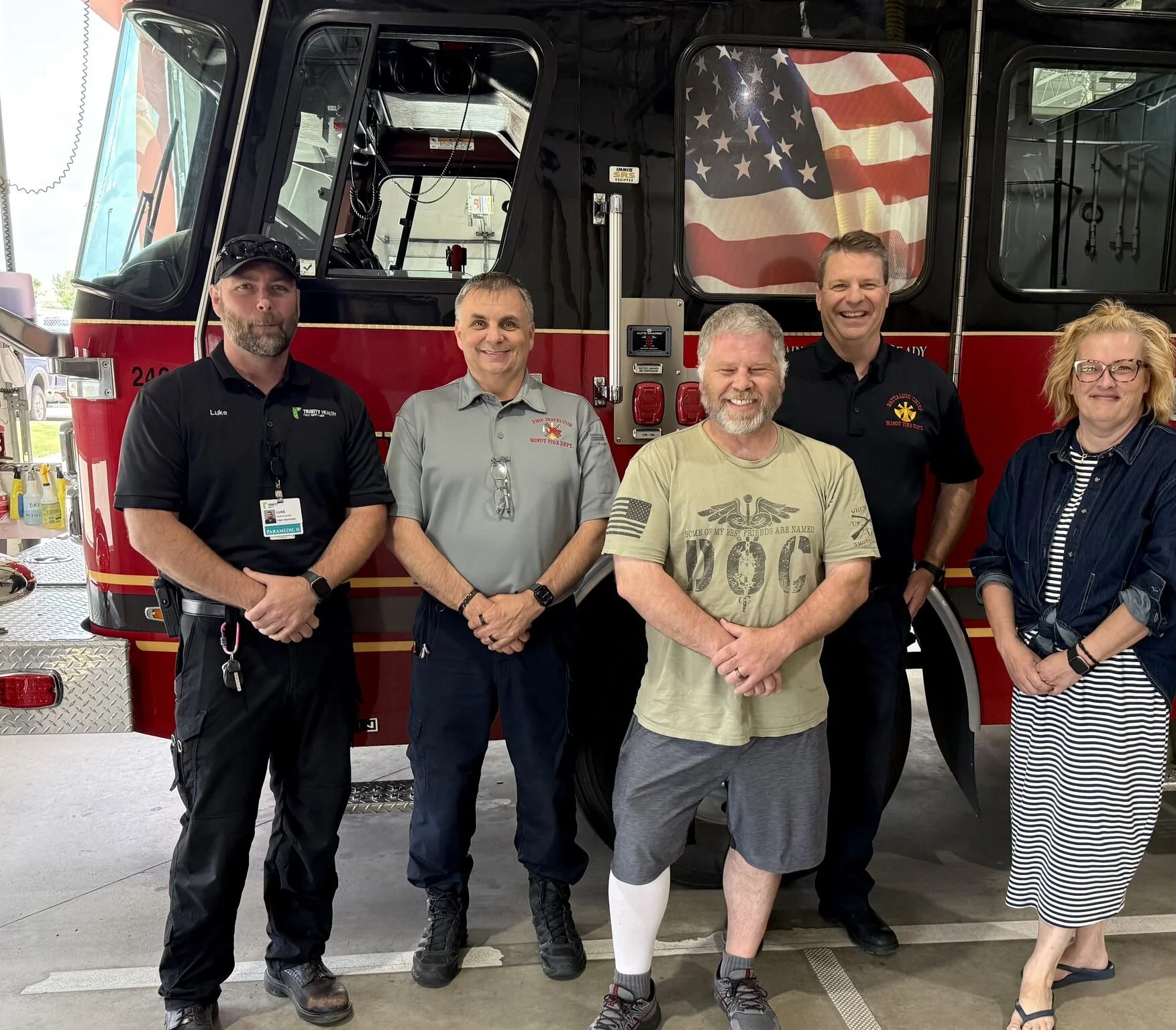 Group of five people standing in front of a fire truck with an American flag window design. Four men and one woman, smiling at the camera, inside a fire station or garage.