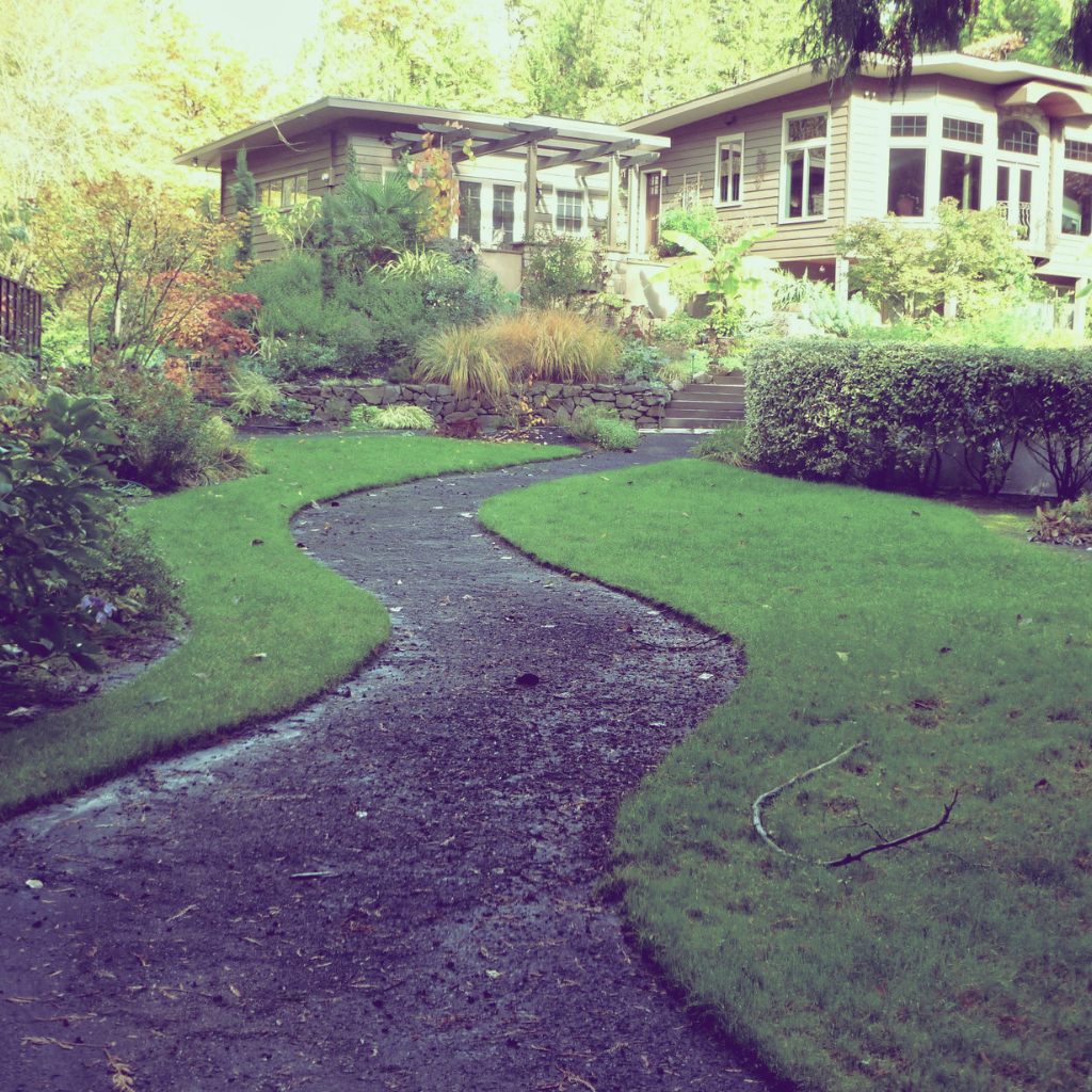 A winding dirt pathway leading to a house with a garden and lush greenery.