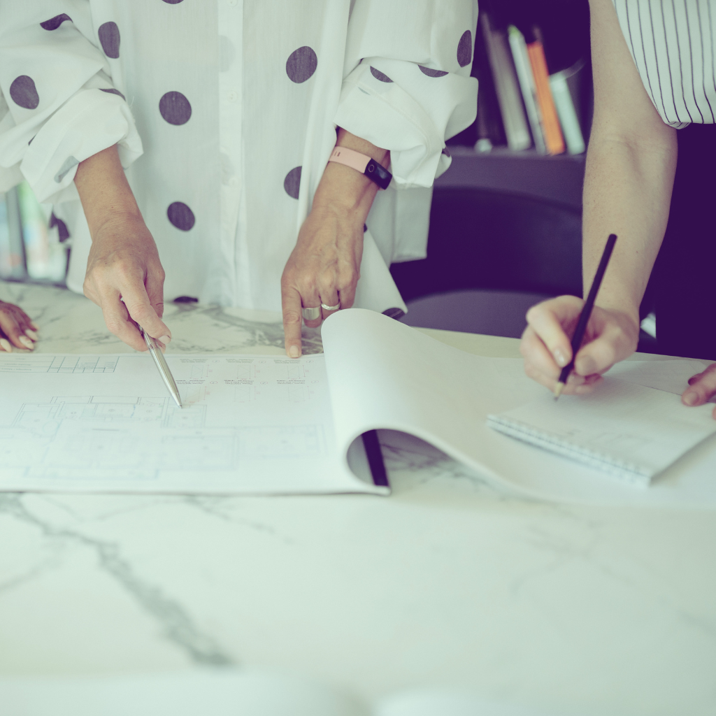Two people reviewing architectural blueprints on a marble table, with one pointing and the other writing notes.