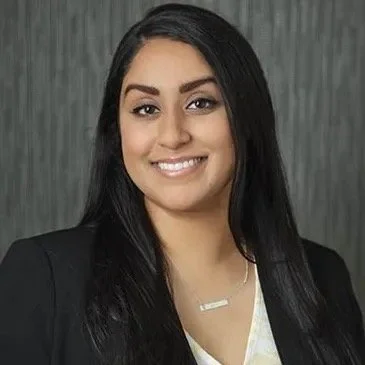 A young woman with long black hair and a bright smile, wearing a black blazer and a white top, standing in front of a gray textured background.