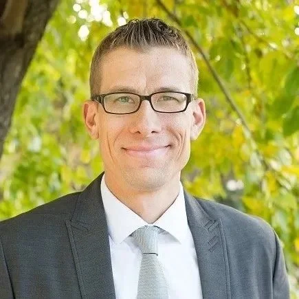 A man in a suit and glasses smiling outdoors with green tree leaves in the background.