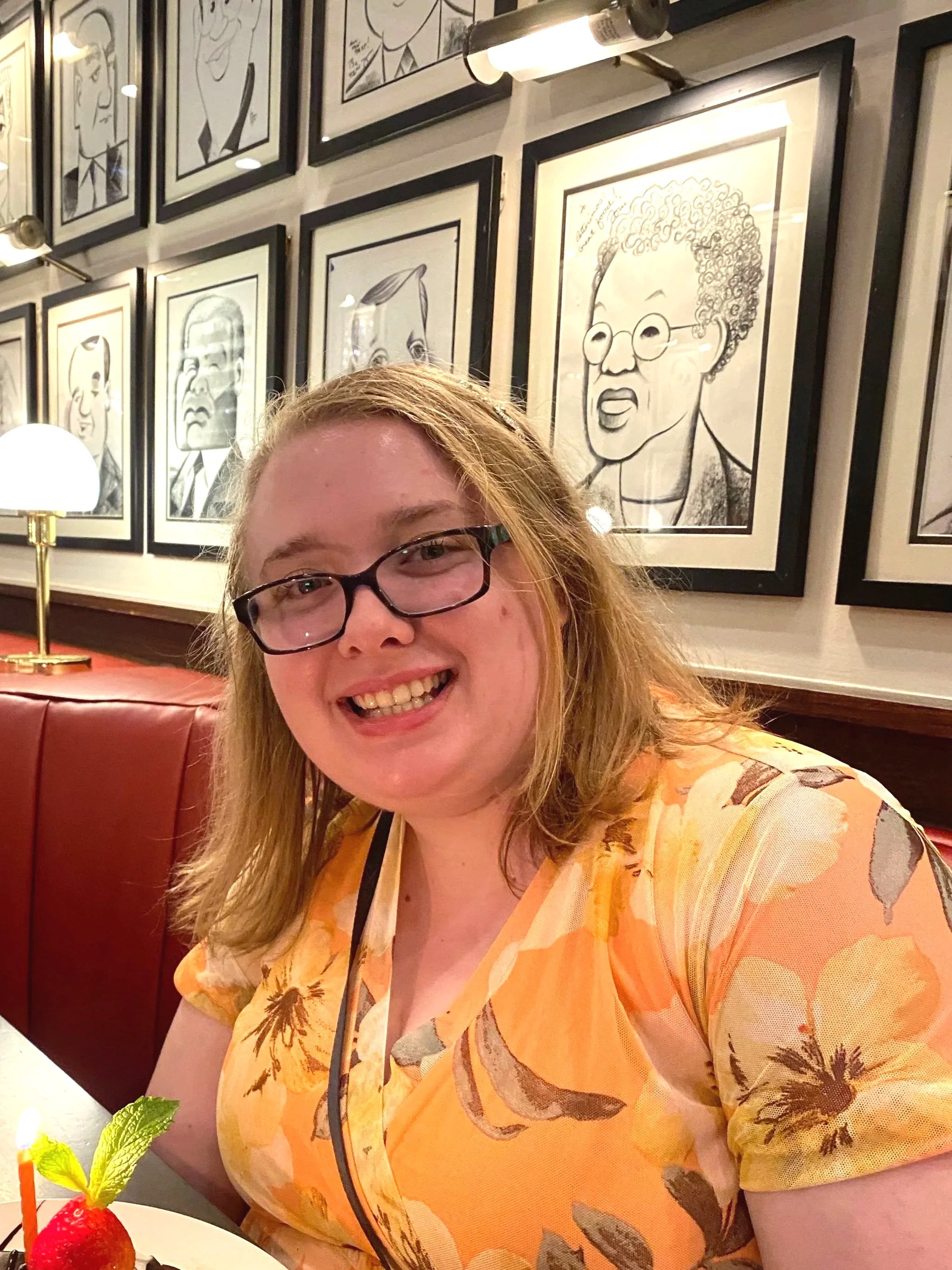 A woman with glasses smiling at the camera, wearing a yellow floral dress, sitting in a restaurant with framed caricature art on the wall behind her, and a white table with a strawberry and a small plant in the foreground.