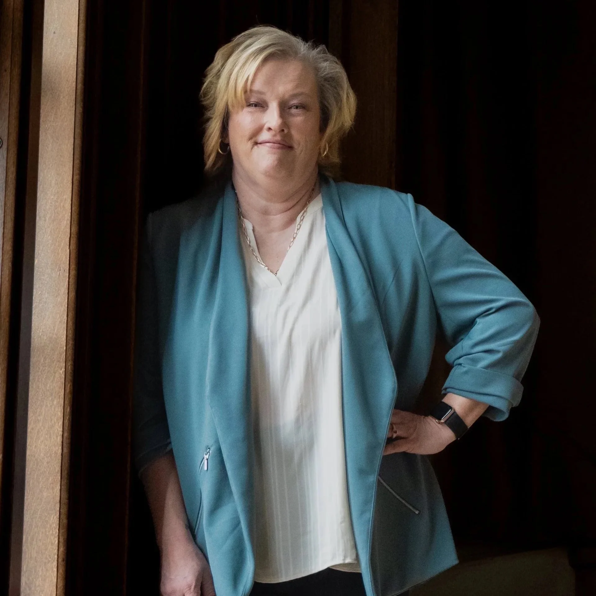 A woman with short blonde hair, wearing a light blue jacket over a white blouse, standing in an elevator with dark walls. She has a slight smile on her face and is wearing a watch and a necklace.
