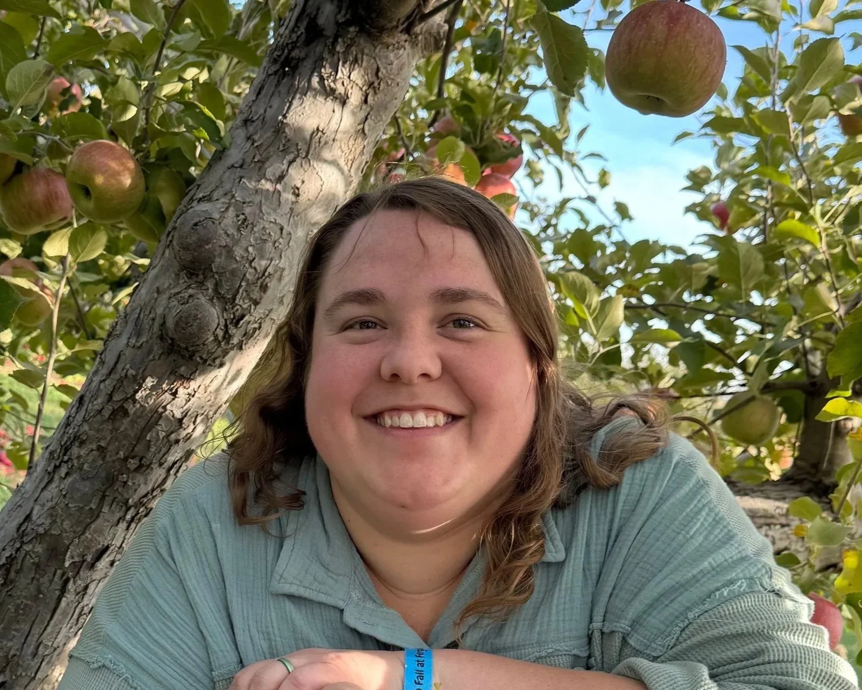 A smiling woman with brown hair taking a selfie outdoors under a tree with green leaves and red apples.