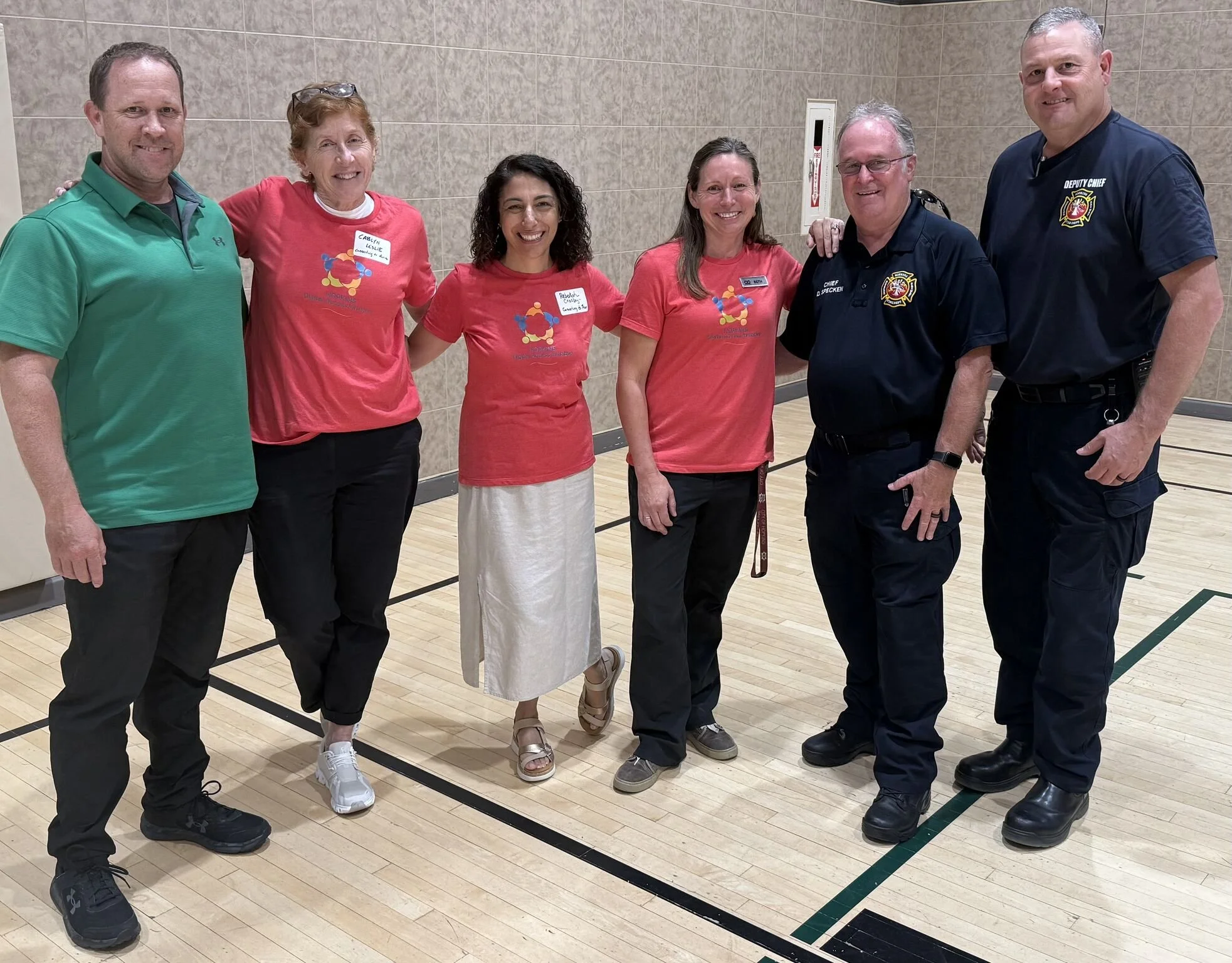 Group of six people standing in a gymnasium, smiling, including firefighters and civilians, some wearing red T-shirts with a logo