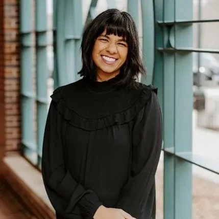 Young woman with shoulder-length dark hair and bangs smiling, wearing a black top with ruffles, standing inside near large windows.