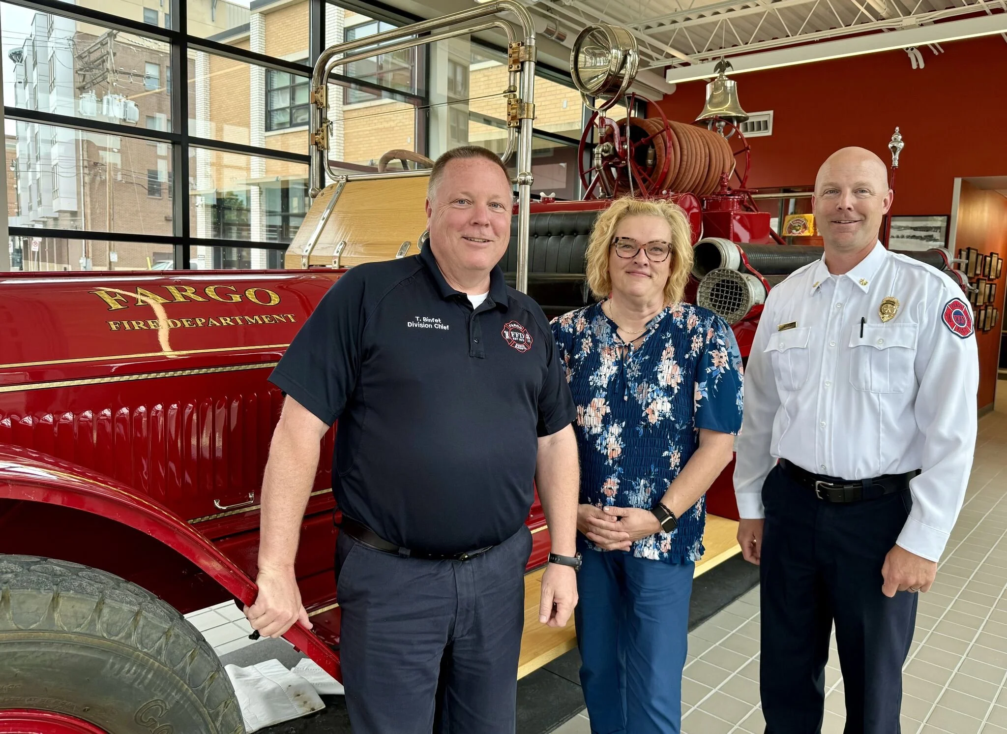Three people standing in front of a vintage red fire truck inside a building with large windows. The fire truck has the words 'FARGO FIRE DEPARTMENT' on its side. The individuals include two men in uniform and a woman in casual attire.