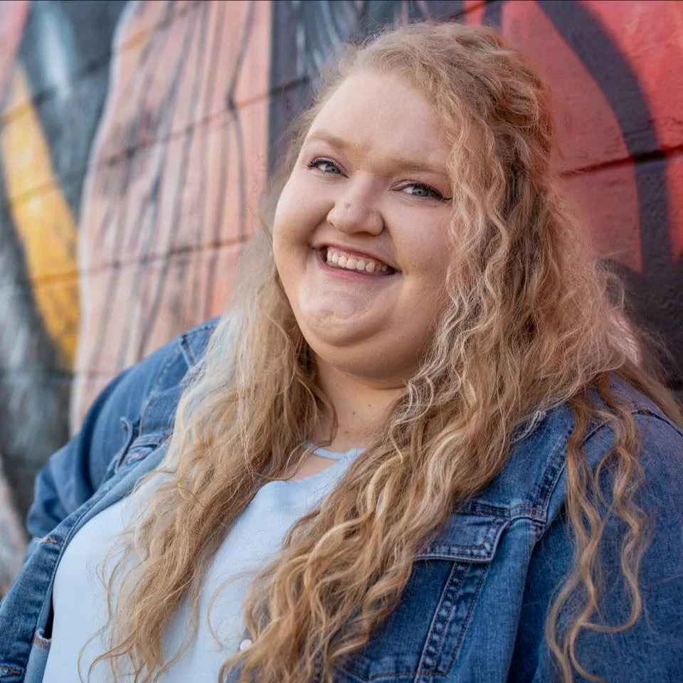 A young woman with long, curly blonde hair smiling in front of a colorful graffiti wall, wearing a denim jacket and a white top.