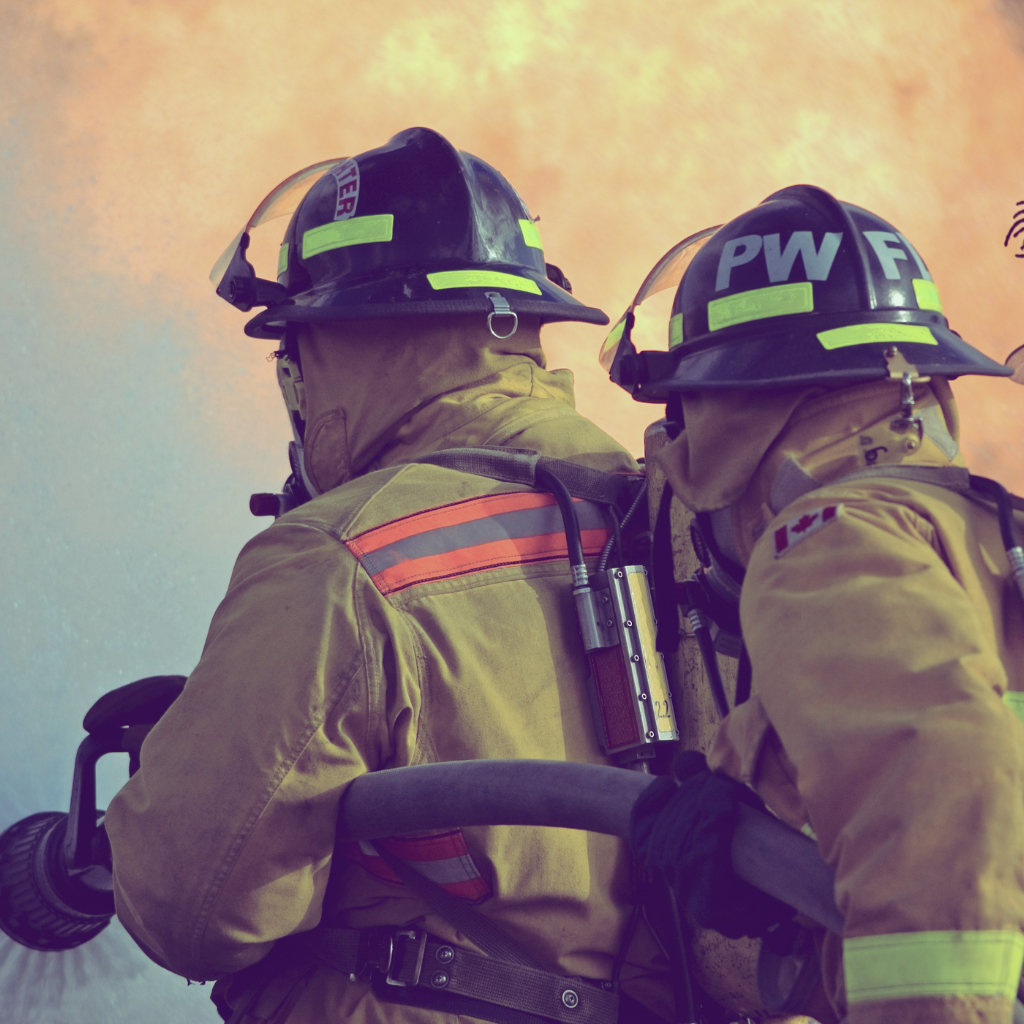 Two firefighters in full protective gear, including helmets and turnout coats, are working together with one holding a hose, against a smoky orange sky.