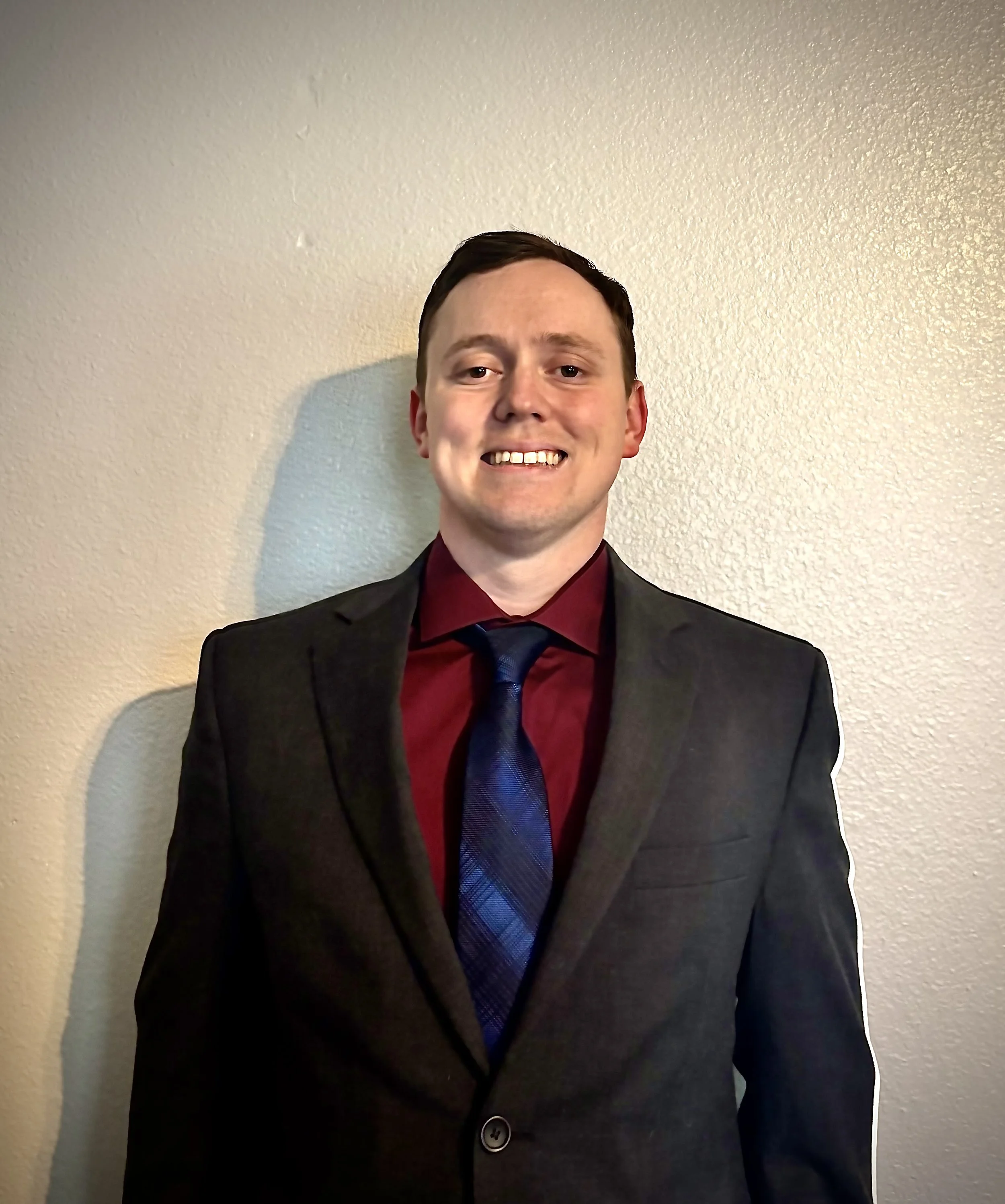 A young man with short dark hair wearing a dark suit, red shirt, and blue tie, smiling against a beige wall.