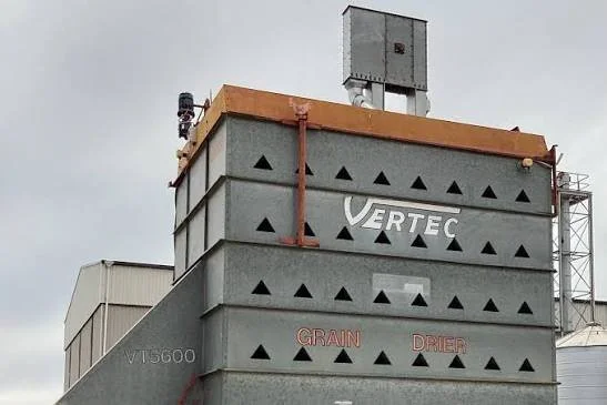 Close-up of a grain dryer structure with the words 'VERTEC' and 'GRAIN DRYER' printed on it, featuring a metal box on top against a cloudy sky.