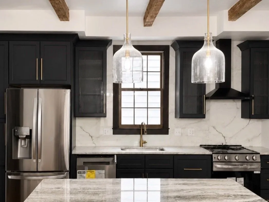 Modern kitchen with dark cabinets, marble backsplash, two glass pendant lights, a stainless steel refrigerator, a gold faucet, and a window above the sink.