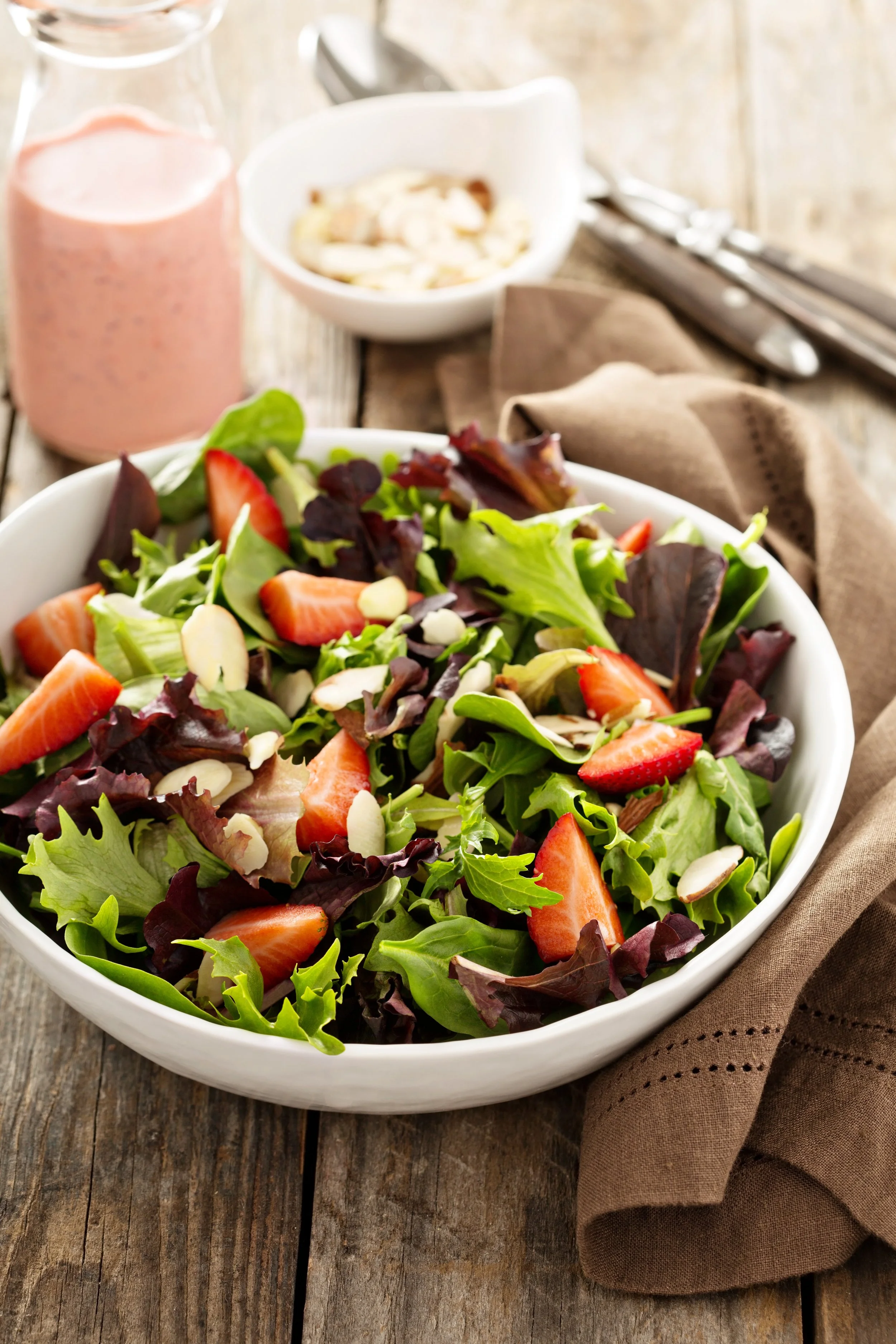 A bowl of fresh salad with strawberries, leafy greens, and sliced almonds on a rustic wooden table.