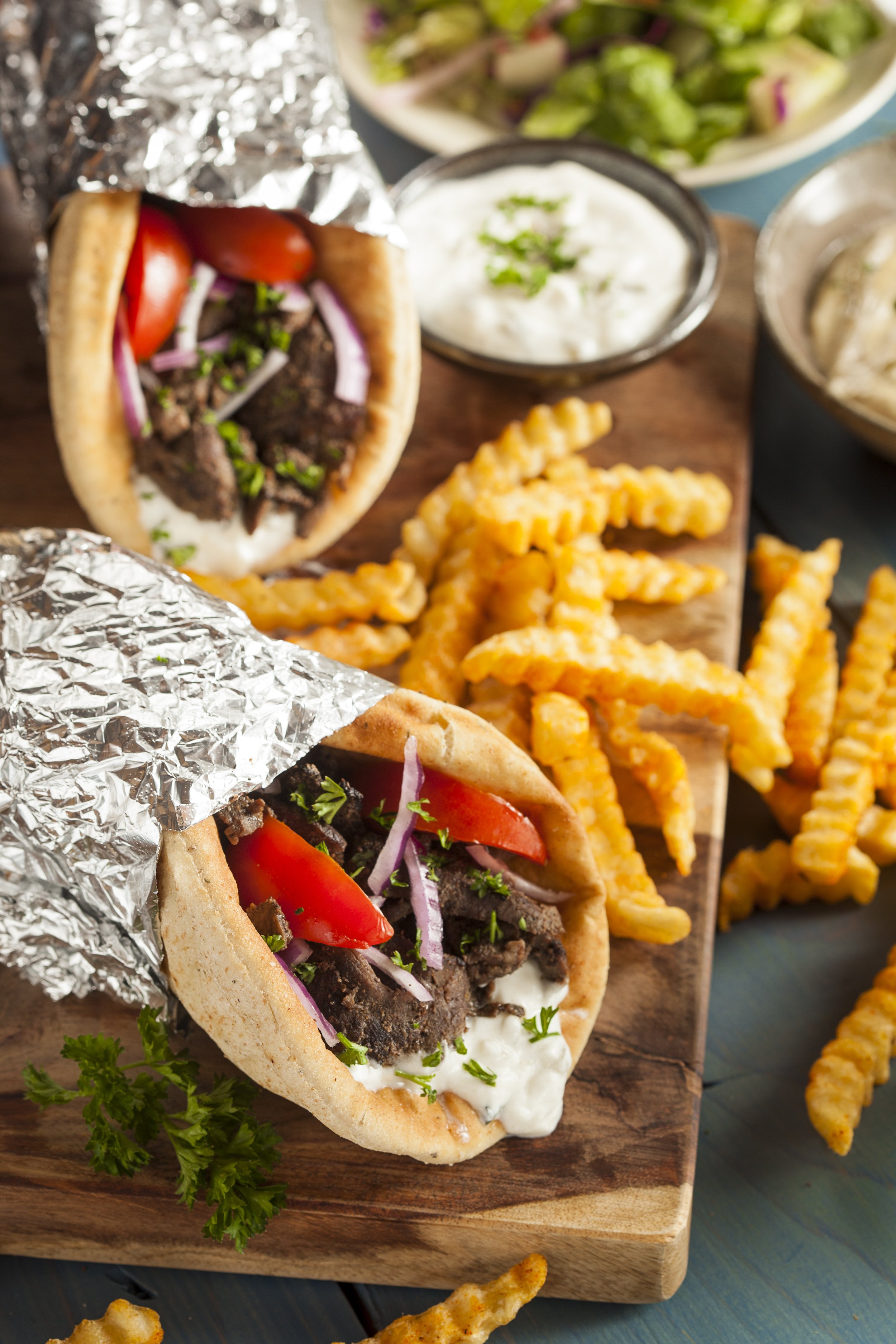 Close-up of two beef shawarma pita sandwiches with toppings, served with crinkle-cut French fries on a wooden board, garnished with parsley. Accompanying bowls of creamy white sauce and green salad are in the background.