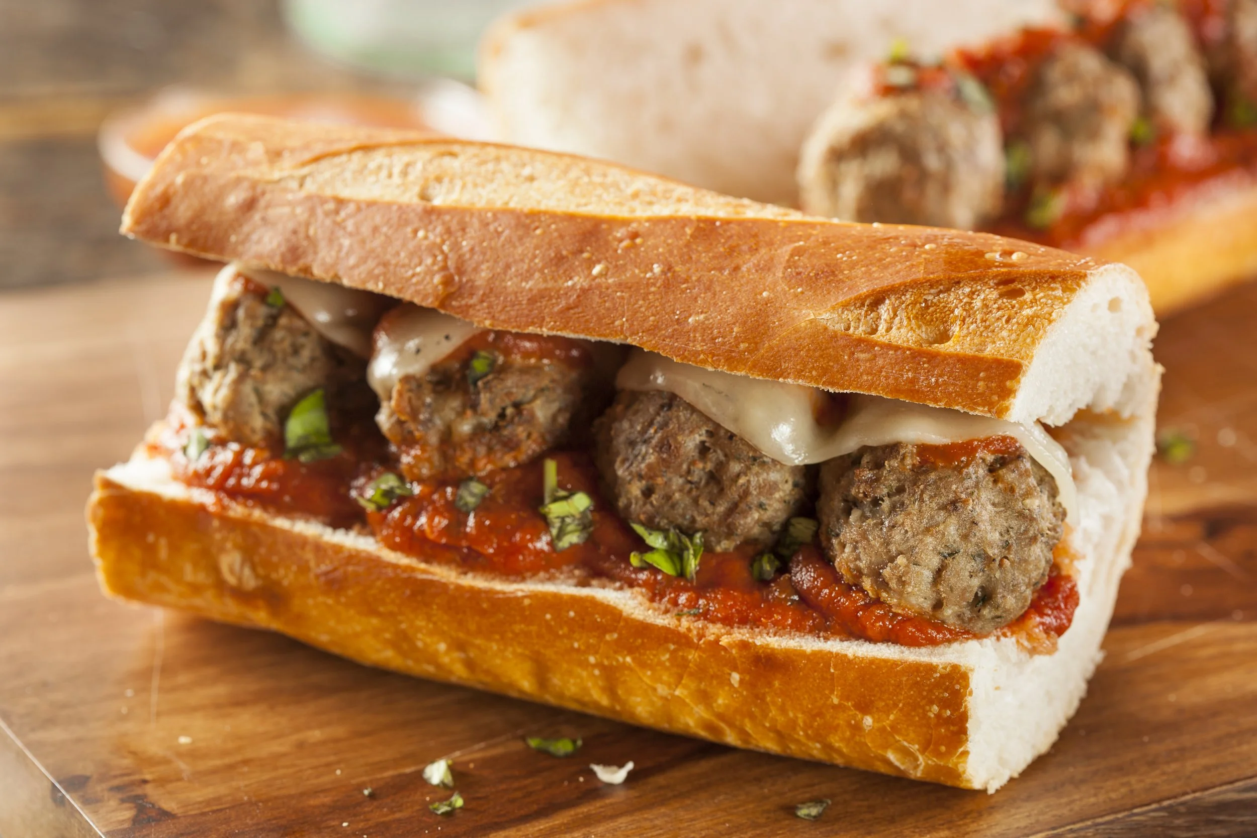 Close-up of a sub sandwich with beef meatballs, melted cheese, marinara sauce, and chopped herbs inside a toasted baguette.