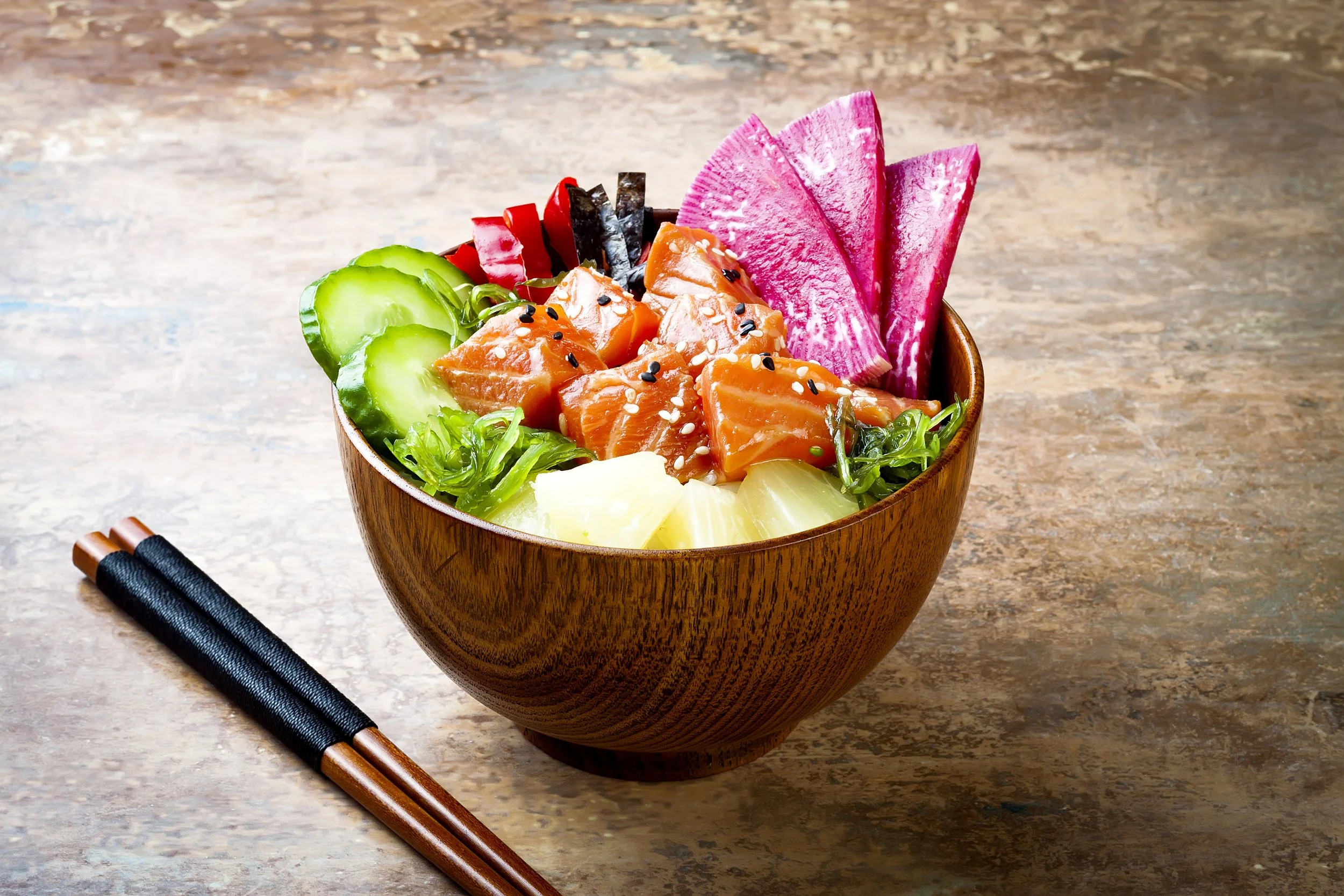 A wooden bowl filled with fresh sliced vegetables and salmon sashimi, garnished with sesame seeds, placed on a wooden surface. Chopsticks are beside the bowl.