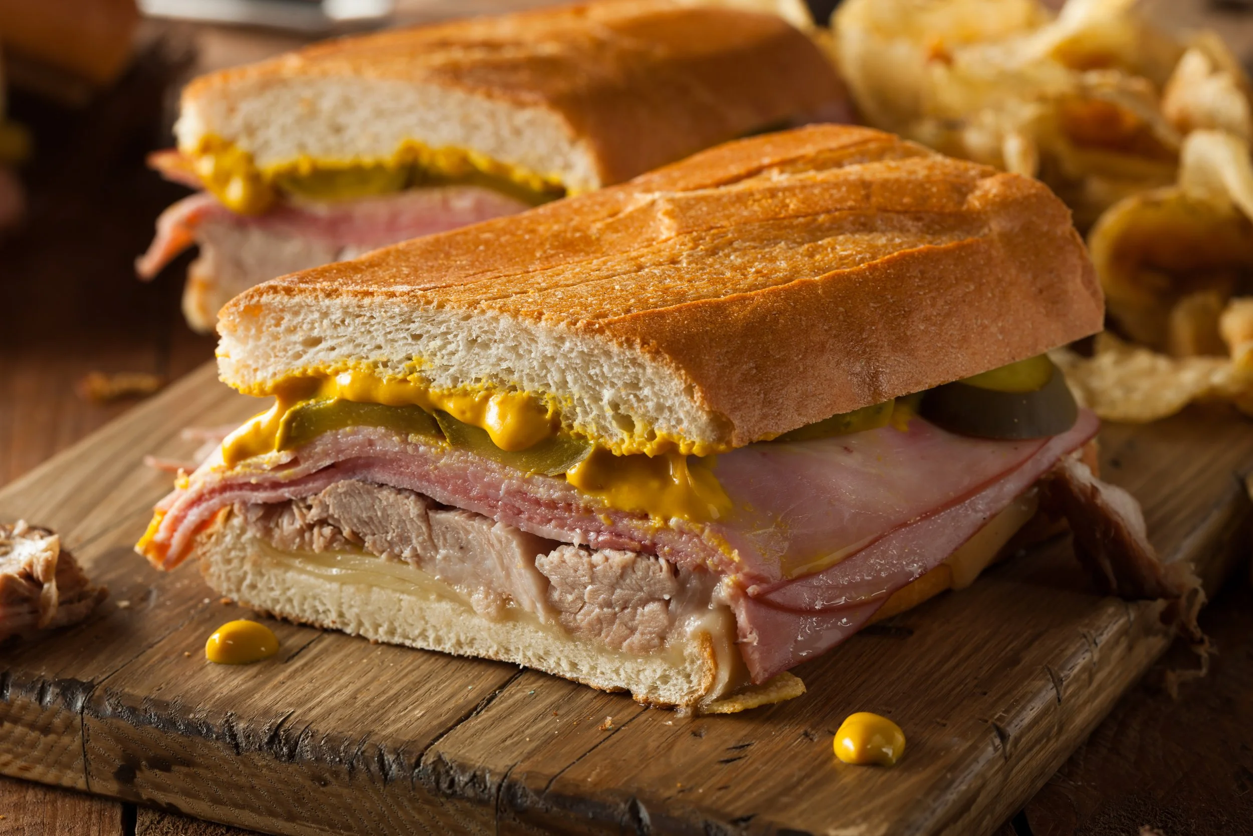 A sandwich on a wooden cutting board with sliced bread, ham, cheese, pickles, and mustard inside.
