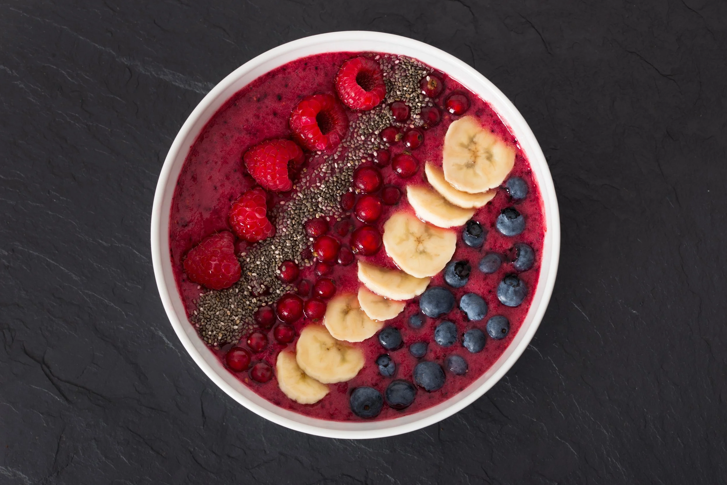 A white bowl filled with a berry smoothie topped with raspberries, banana slices, blueberries, red currants, chia seeds, and hemp seeds, placed on a black textured surface.