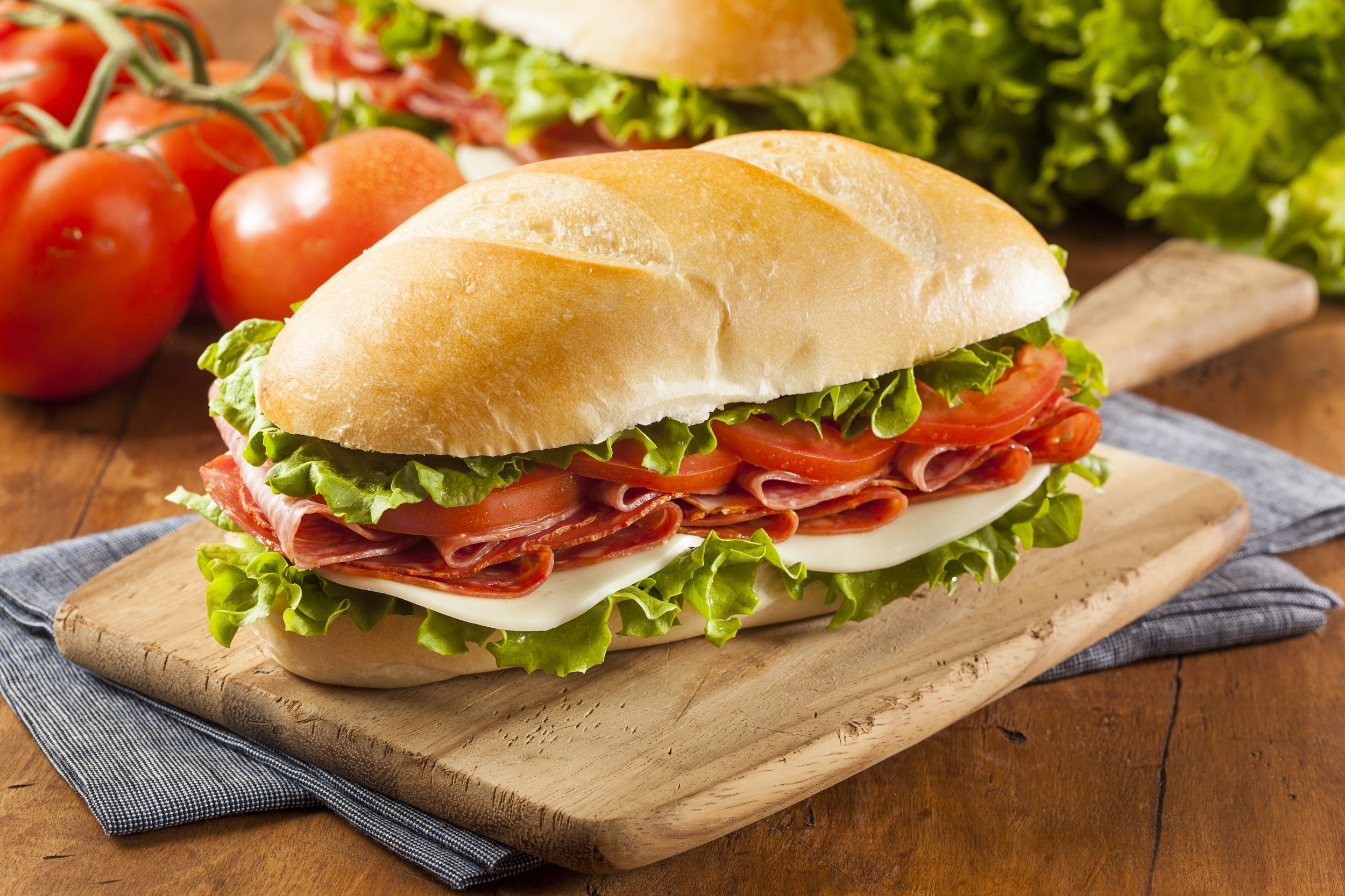 A sandwich with lettuce, tomato, cheese, and sliced meat on a wooden cutting board, with tomatoes and leafy greens in the background.