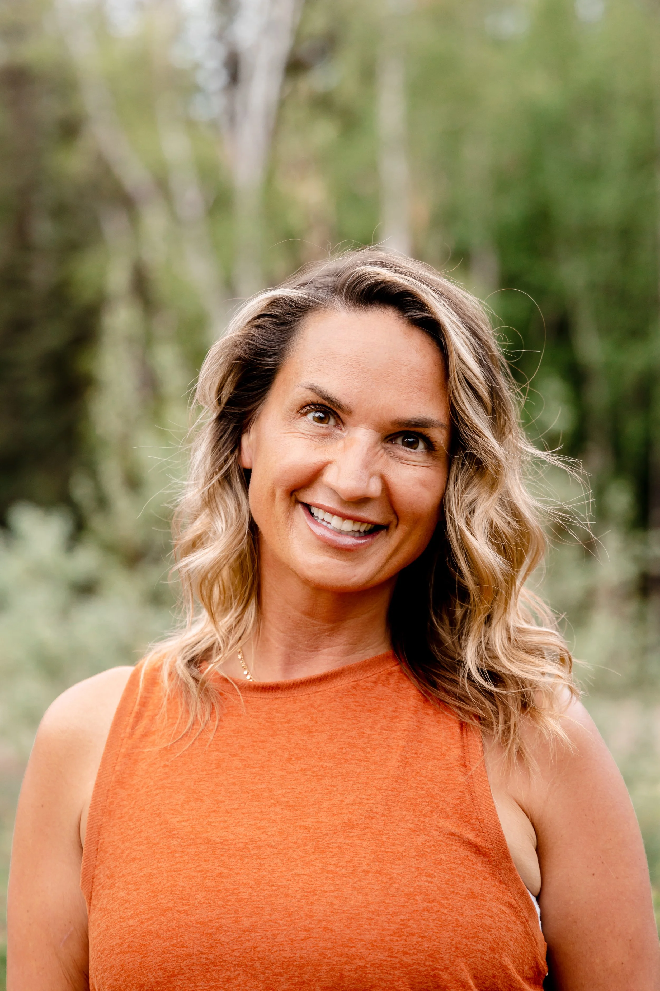 Close-up of a smiling woman with wavy blonde hair, wearing an orange sleeveless top, outdoors with blurred green trees in the background.