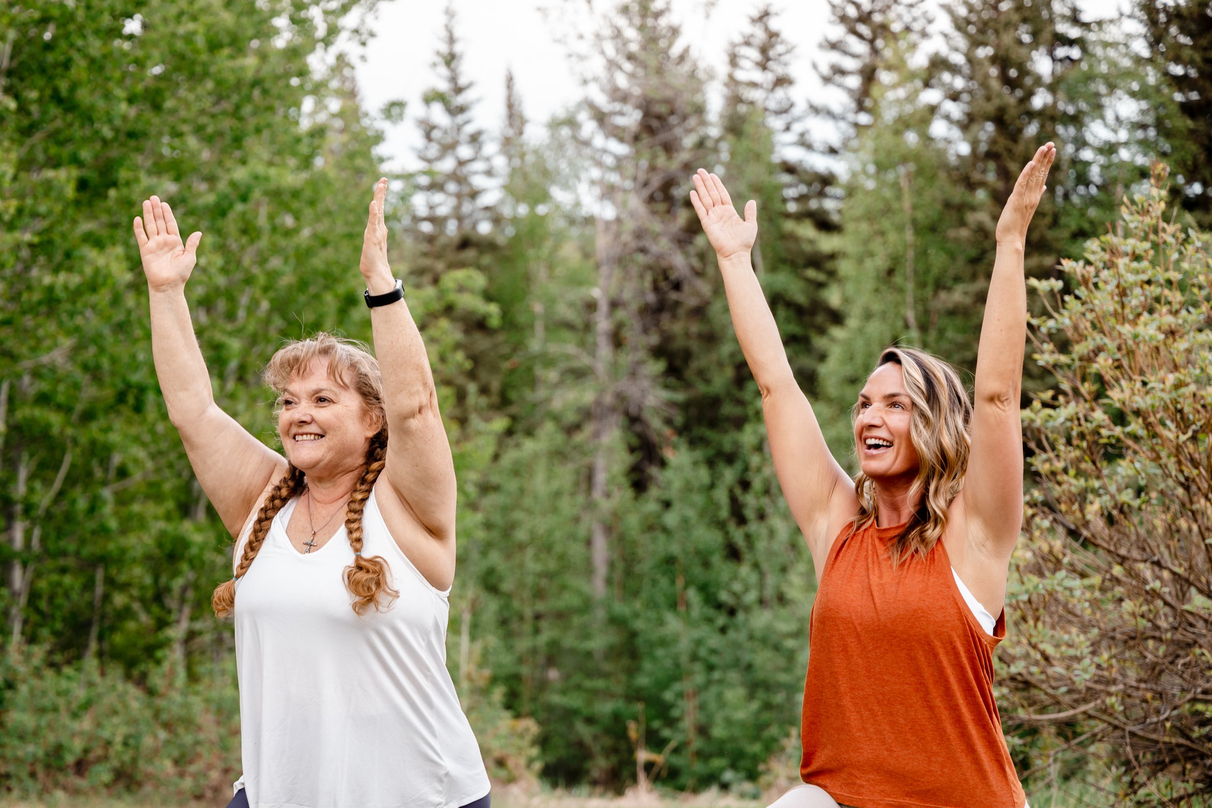 Two women exercising outdoors, raising their arms, with trees in the background.