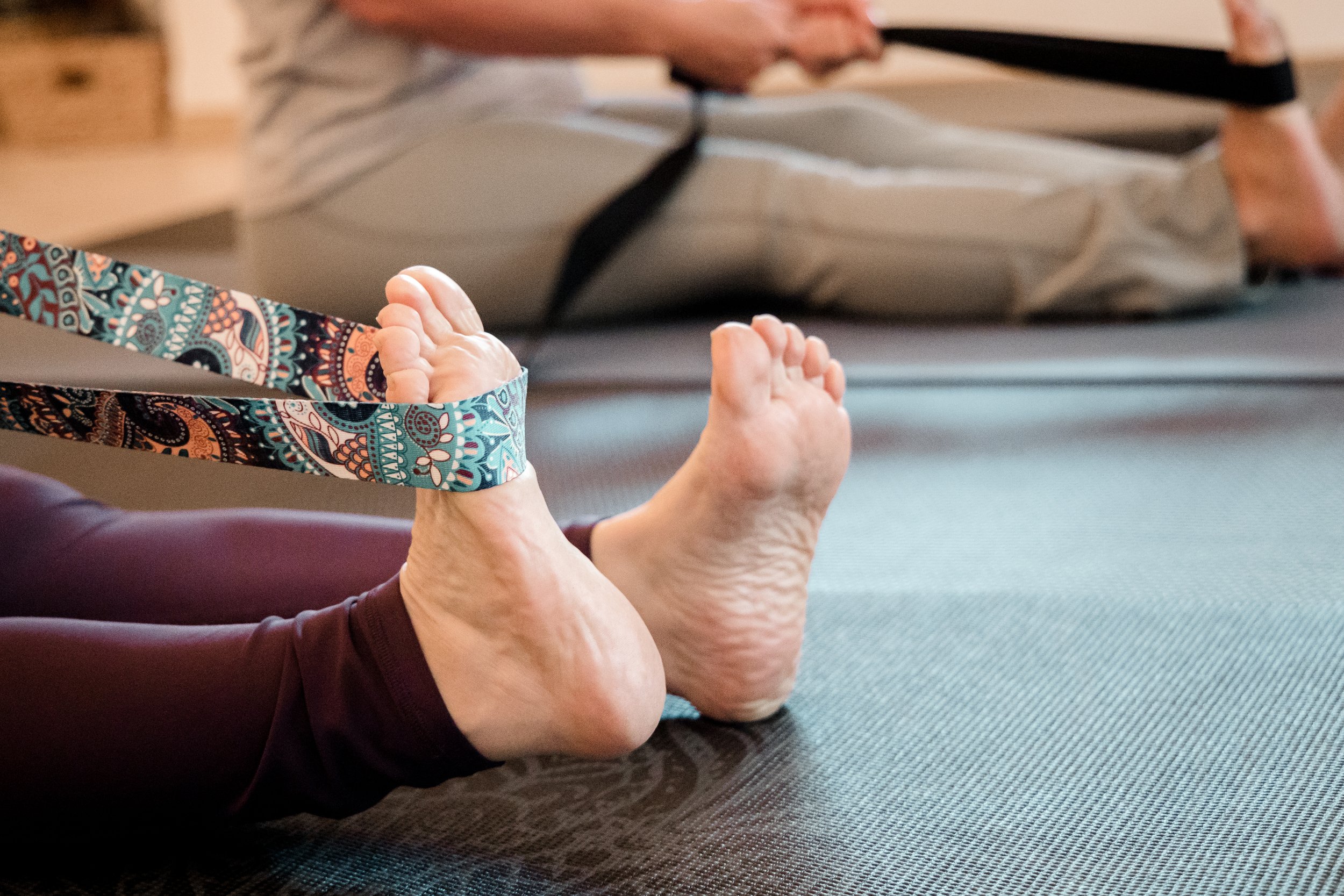 A person lying on their stomach on a yoga mat, with their feet visible and a decorative fabric strap around their ankles, participating in a seated forward bend yoga pose during a class.
