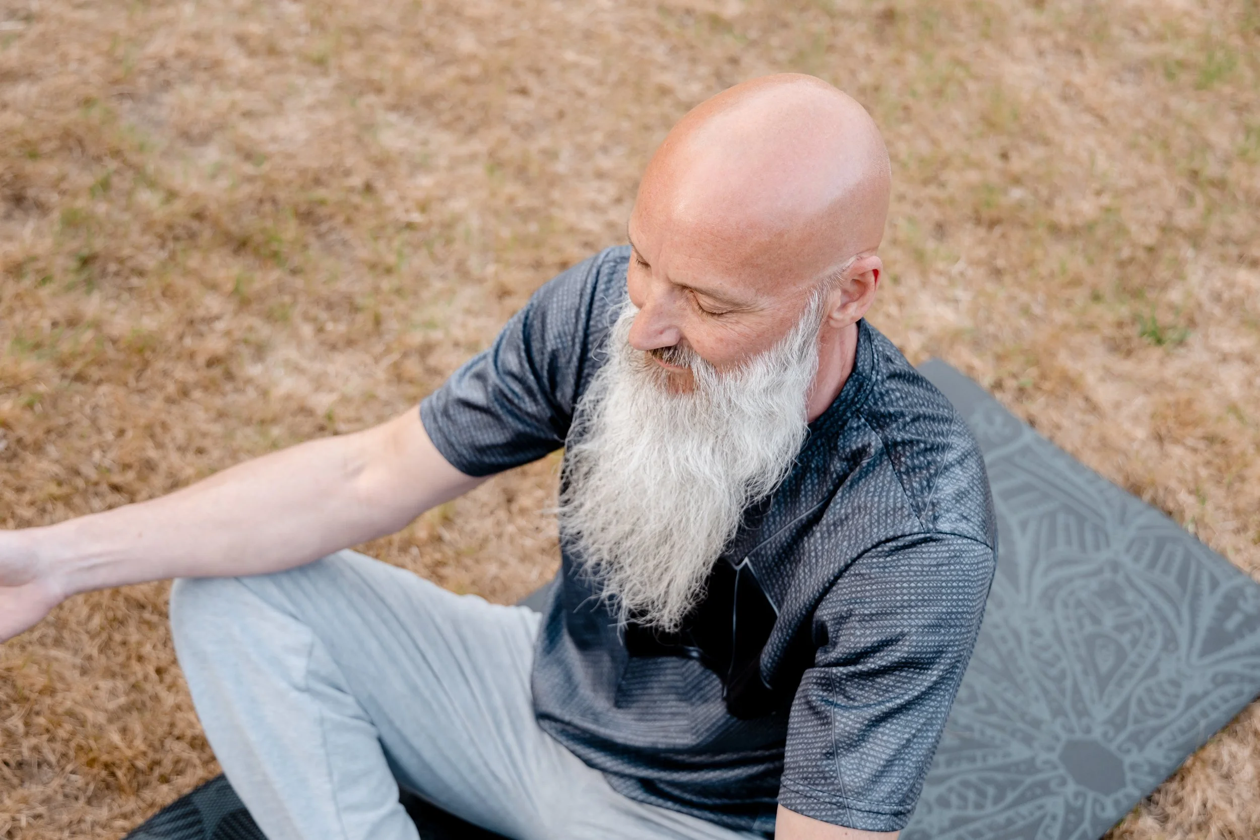 An elderly man with a bald head and long white beard practicing yoga outdoors on a mat on a grassy field.