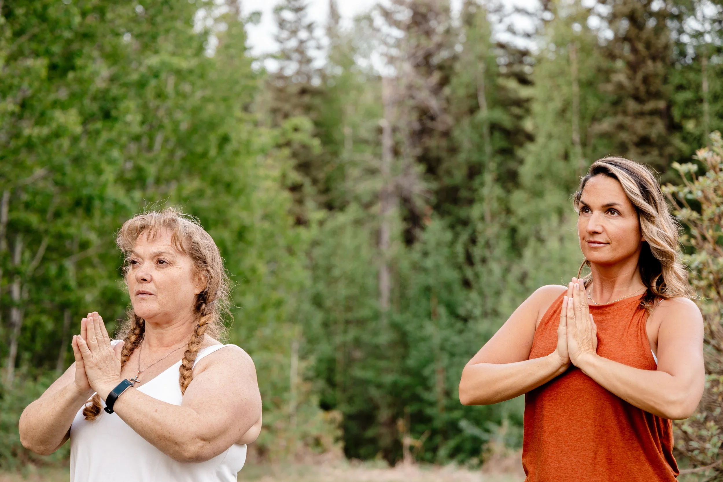 Two women practicing yoga outdoors in a forested area, standing with their hands in prayer position and eyes closed.