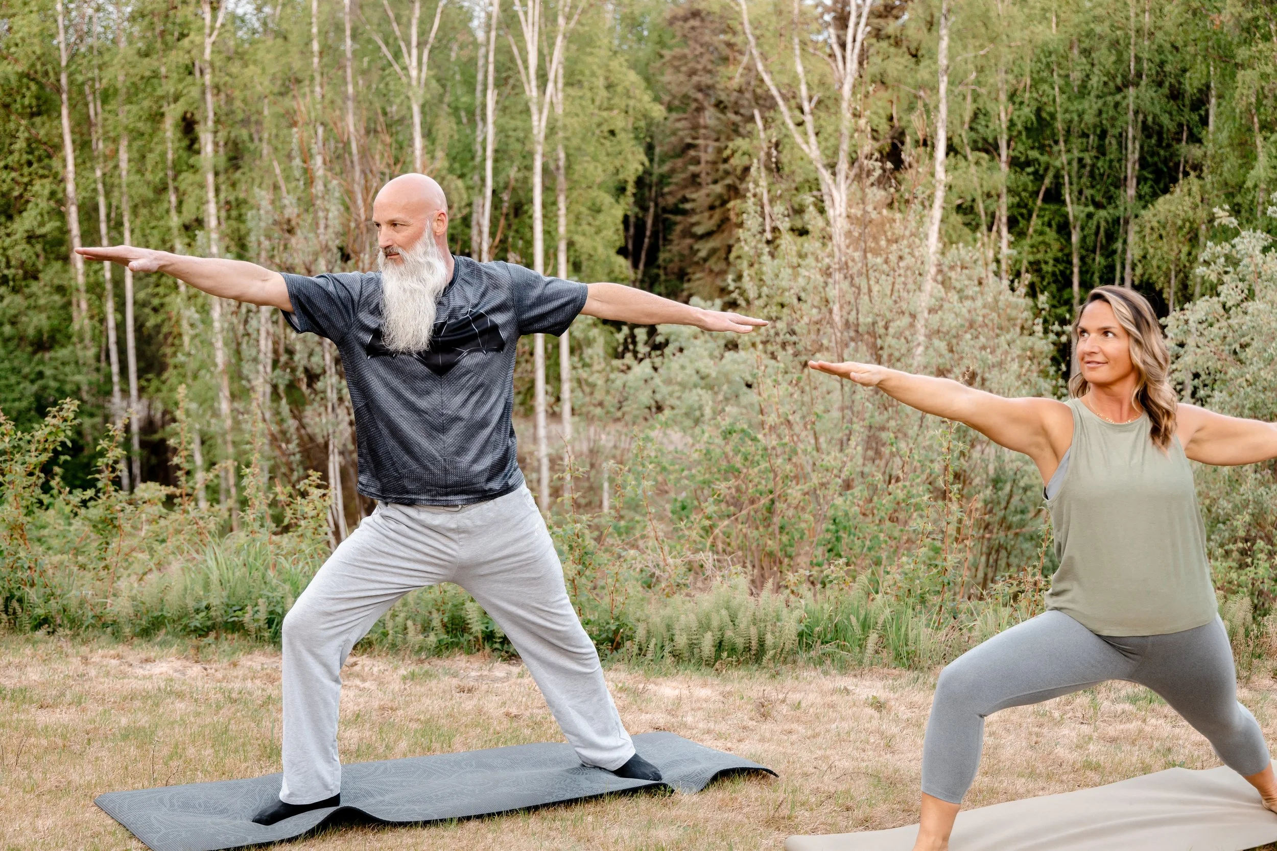 Two people practicing yoga outdoors in a forest, with one person in a Warrior pose and the other person in a Warrior pose, both on yoga mats.
