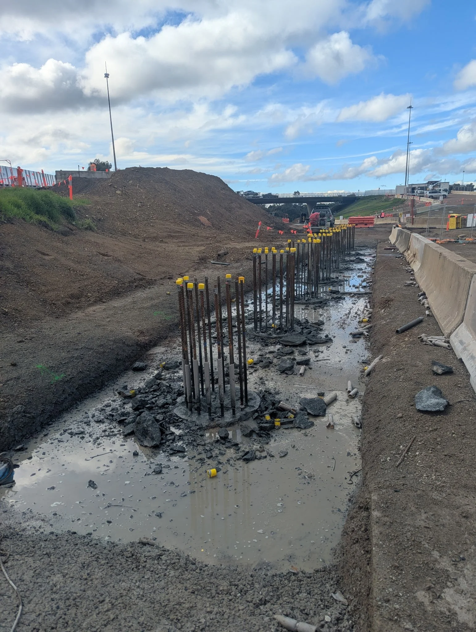 Construction site with exposed rebar and muddy water, surrounded by dirt mounds, fencing, and parked vehicles under a cloudy sky.