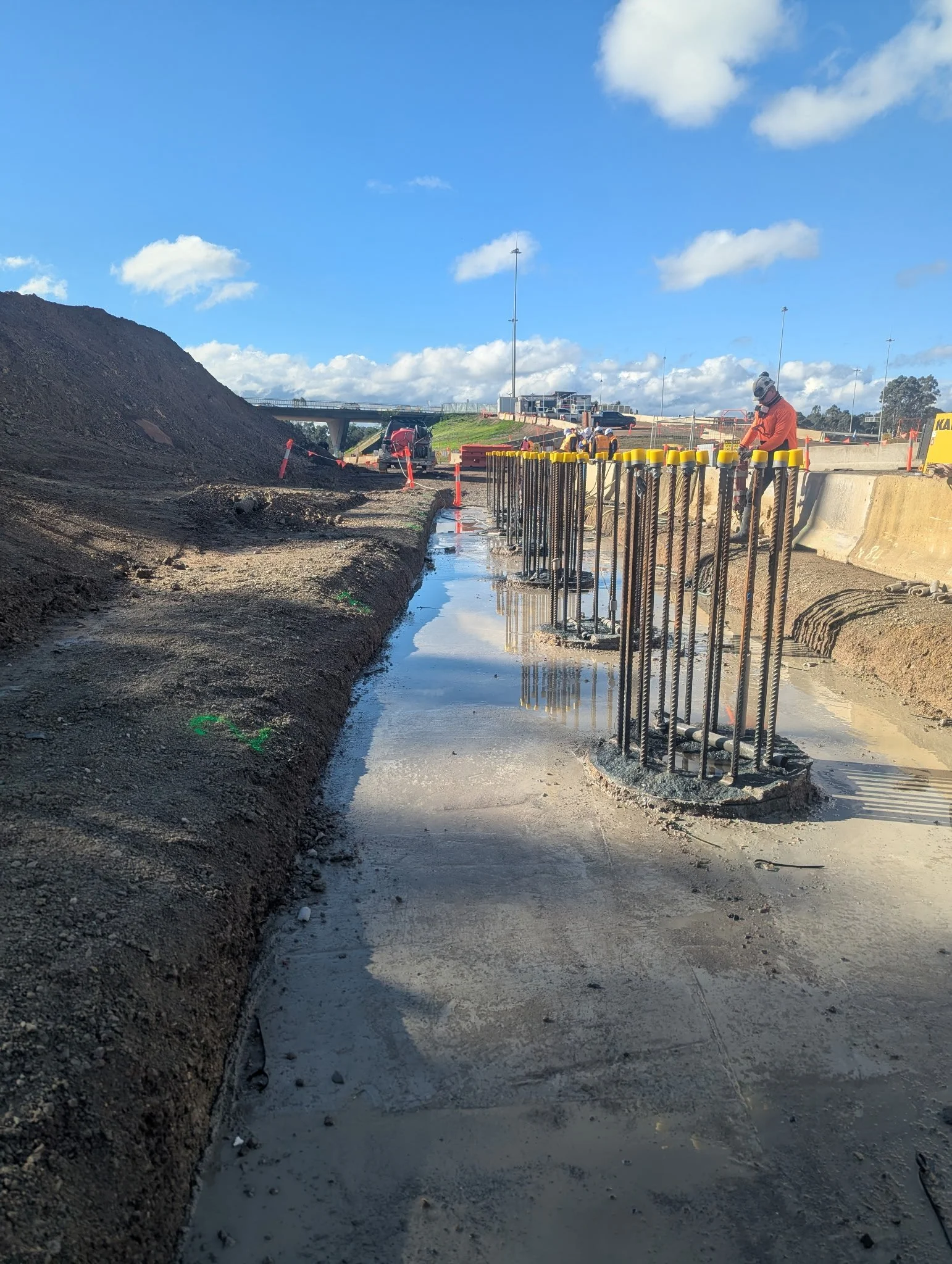 Construction workers on a road project with rebar and concrete, dirt piles, and a bridge in the background under a partly cloudy sky.