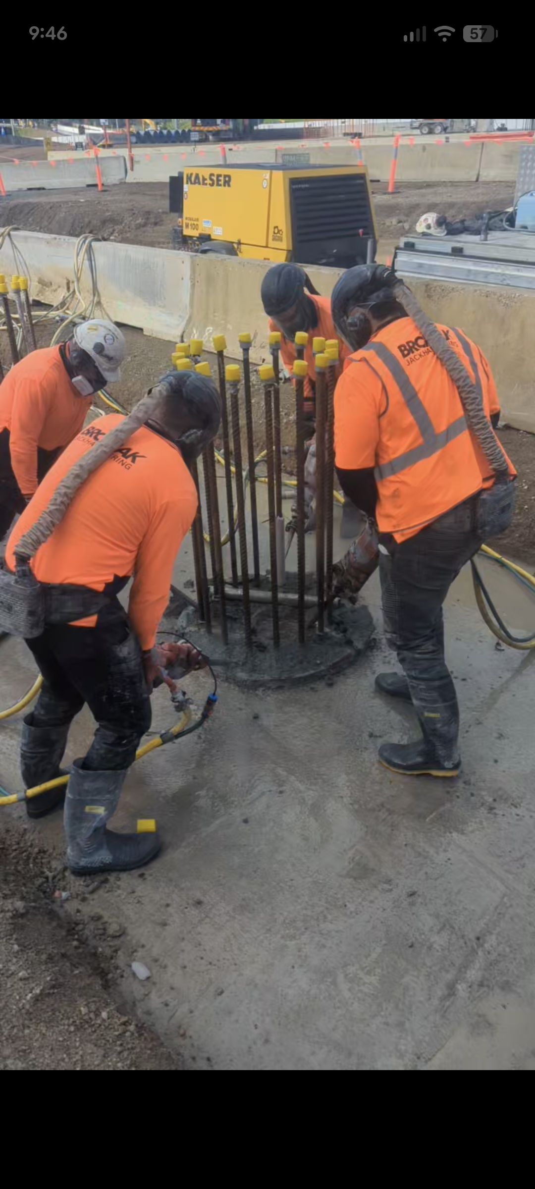 Construction workers in orange safety clothing and helmets working around a metal rebar structure at a construction site.