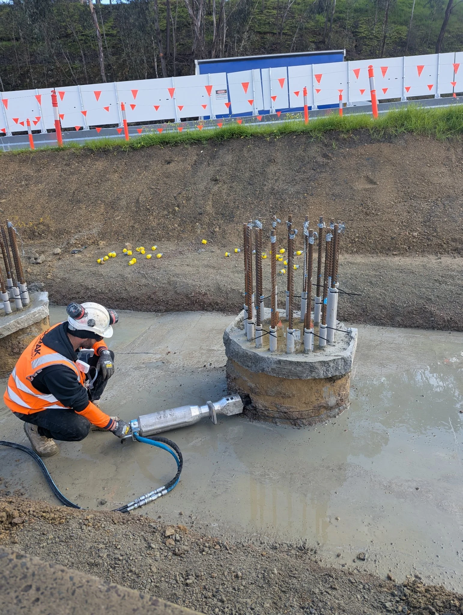 Construction worker in an orange safety vest and white helmet operating equipment on a concrete foundation at a construction site. Steel reinforcement bars are visible in the background, along with a large circular concrete base and a pipe connected to the system.