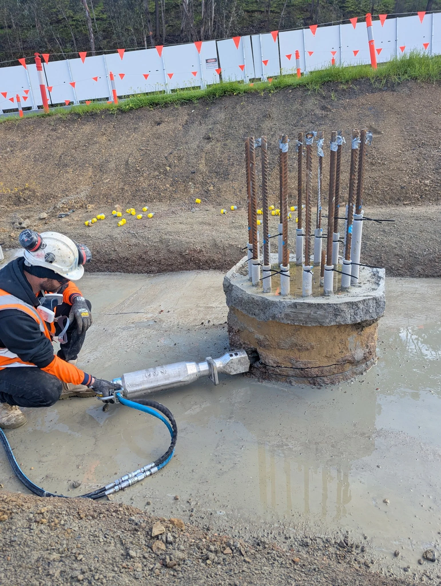 A construction worker wearing a white helmet and orange safety vest working on a concrete and rebar foundation. There are orange safety barriers and warning signs behind him, and a patch of grass and trees in the background.
