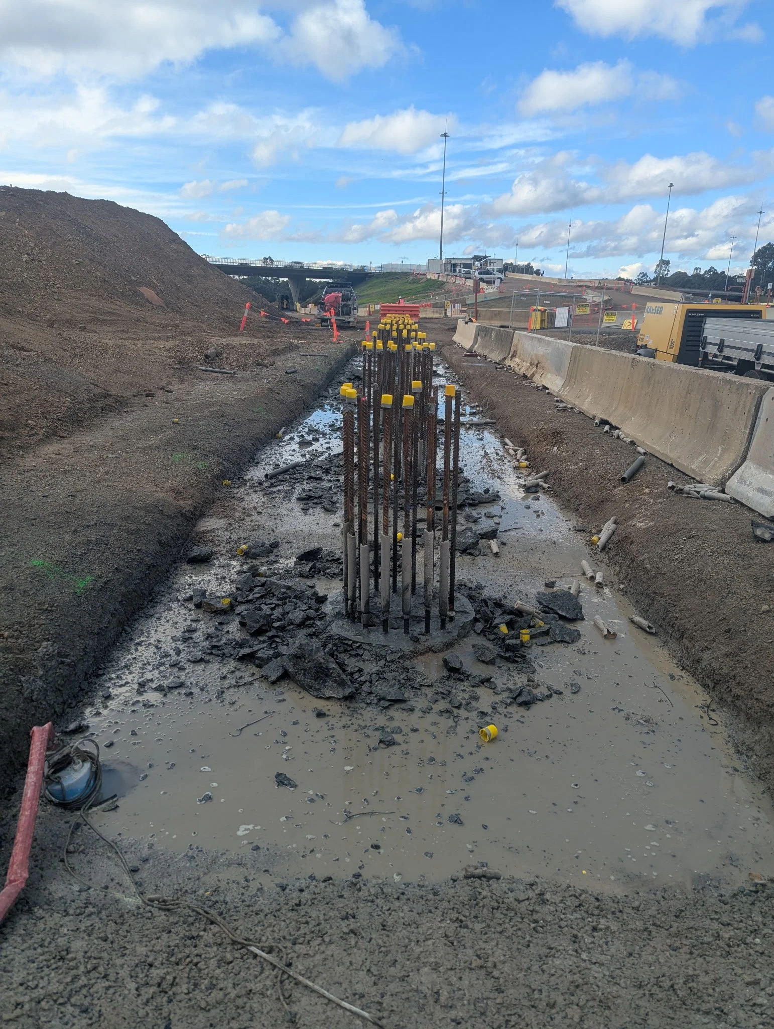 Construction site with exposed rebar in wet concrete, barriers, machinery, and a clear sky with scattered clouds.