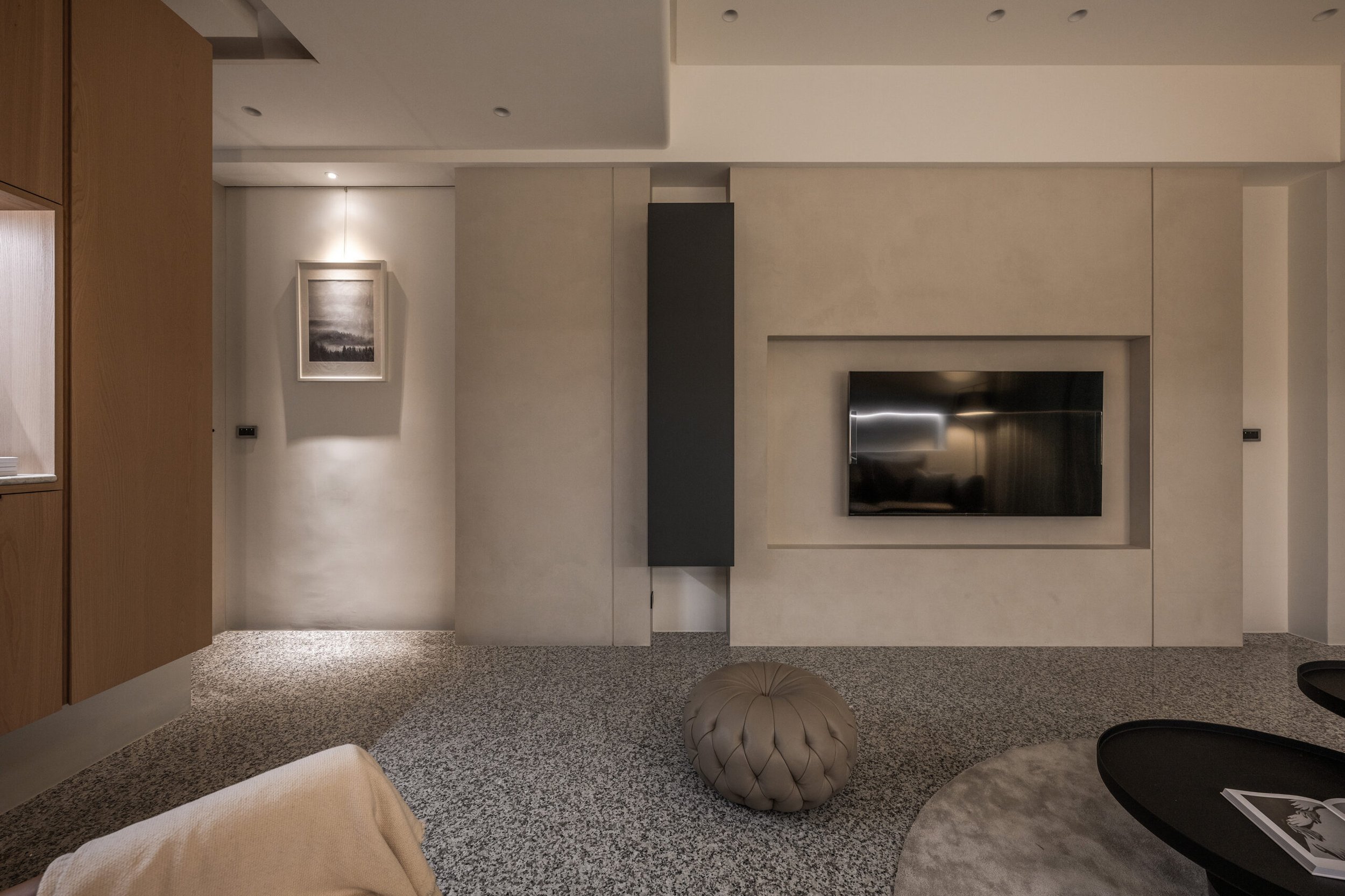 Modern living room with wall-mounted flat-screen TV, beige sofa, round tufted ottoman, artwork, and textured carpet.