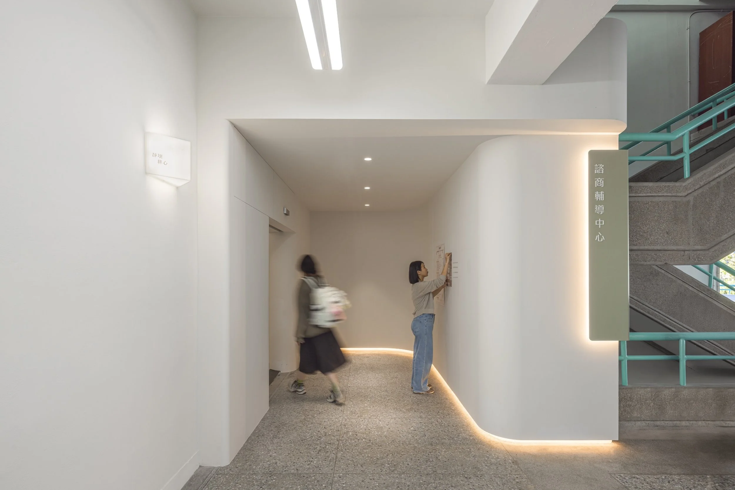 Two women standing in a modern, well-lit interior hallway with white walls and an illuminated sign with Chinese characters. One woman is reading a notice on the wall while the other walks by with a backpack. There is a staircase with teal railings on the right side of the image.