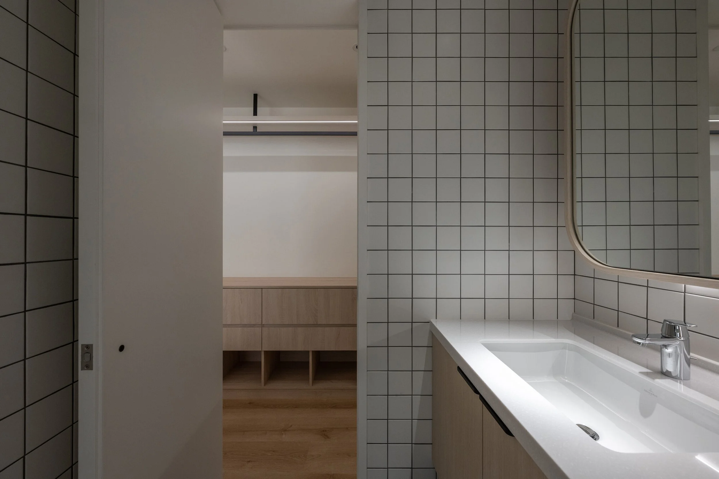 Modern bathroom with white tiled walls, a white sink with a chrome faucet, a large mirror, and wooden cabinets.