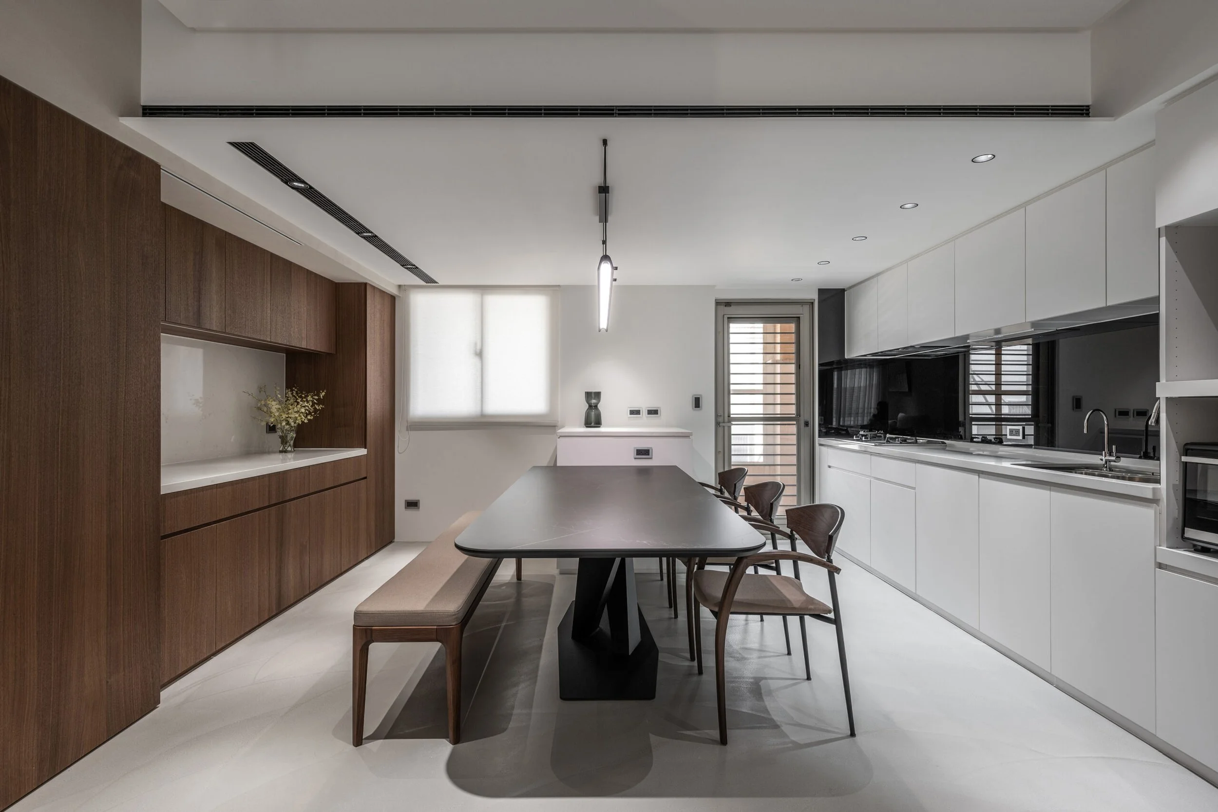 Modern kitchen with white cabinets, black countertop, wooden wall panel, dining table with chairs, and minimalistic lighting.