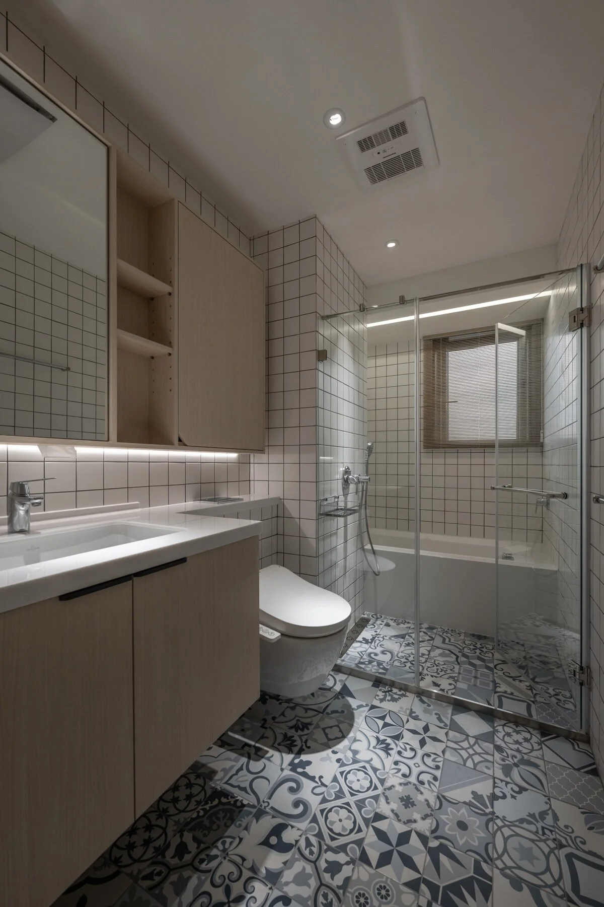 Modern bathroom with patterned floor tiles, white sink, wooden cabinet, toilet, shower with glass doors, and a window with blinds.