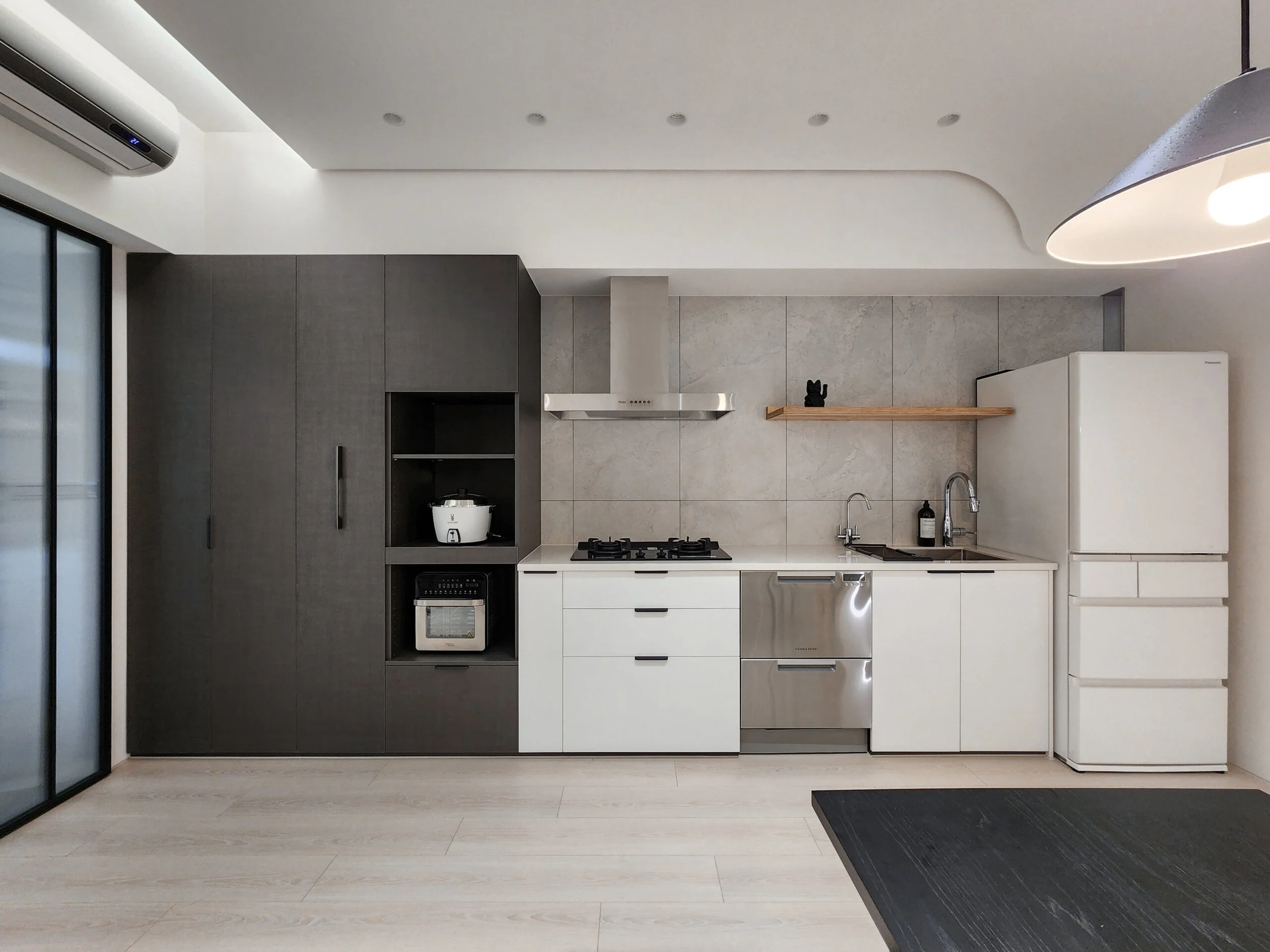 Modern kitchen with white and gray cabinetry, a gas stove, a hood, a small refrigerator, and a black countertop. There is a shelf with a black decorative item, a rice cooker, and a black mat on the light wooden floor.
