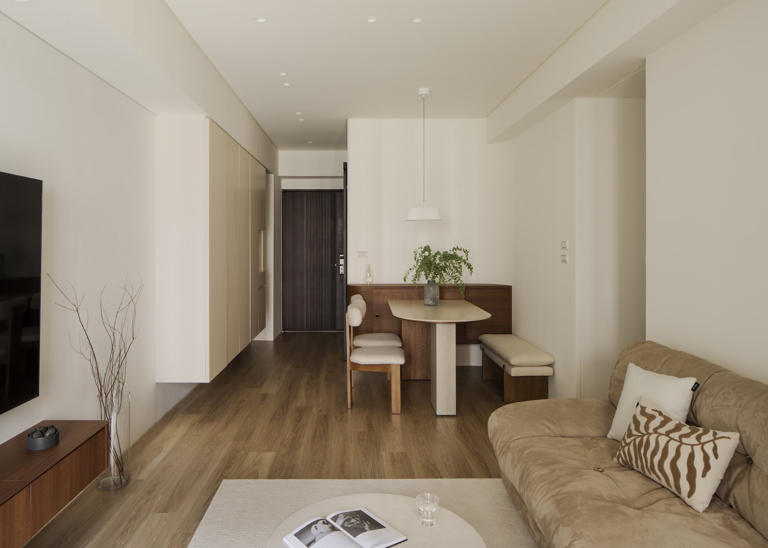 Living room with beige sofa, pillows, coffee table with open magazine and glass of water, wooden flooring, a beige armchair, minimalist decoration, and a dining area with a wooden table and chairs, natural light