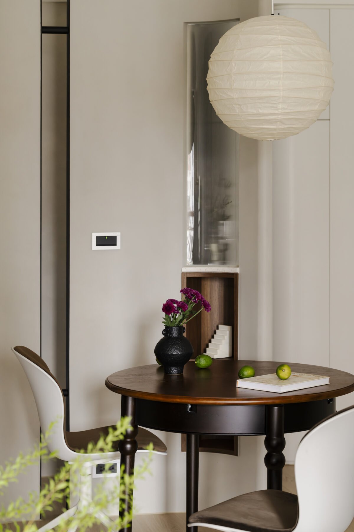 Interior dining area with a round wooden table, white and brown chairs, a ceiling paper lantern, a black vase with pink flowers, green apples, and a small wall shelf with decorative items.