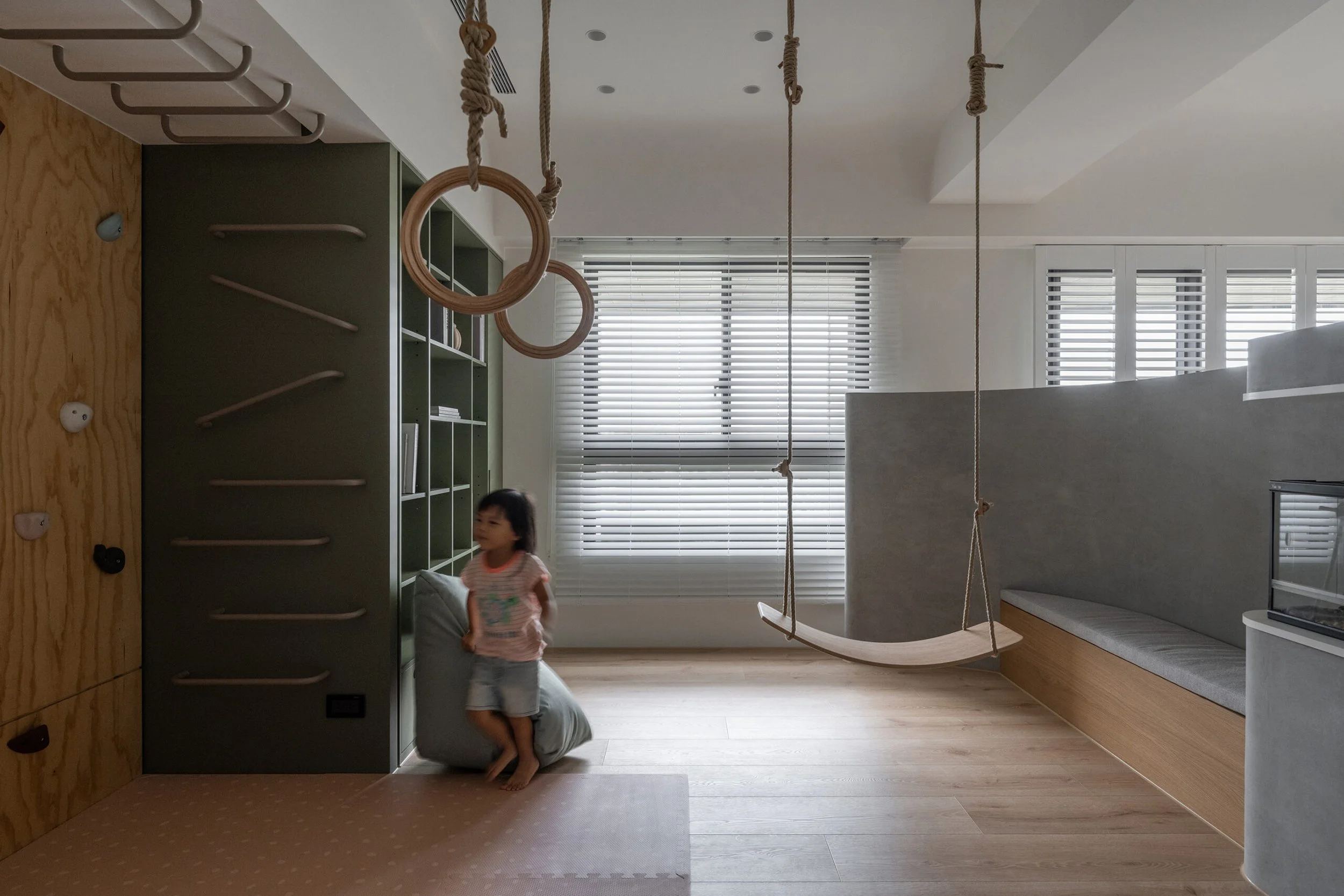 A young girl wearing pajamas standing on a bean bag in a modern playroom with gymnastic rings, hanging ropes, a climbing wall, and large windows with blinds.