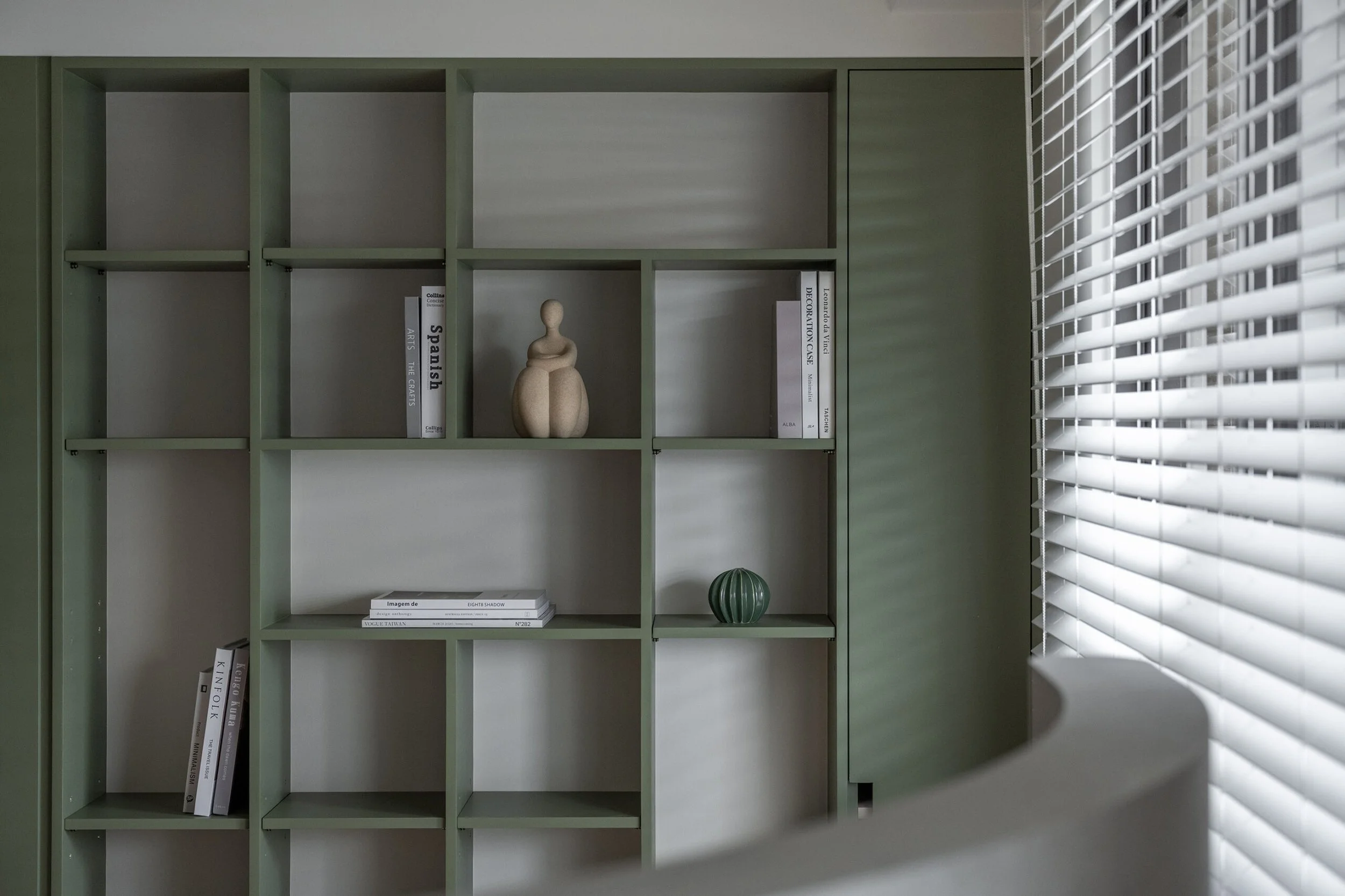 Green bookshelf with books and decorative items near a window with white blinds.