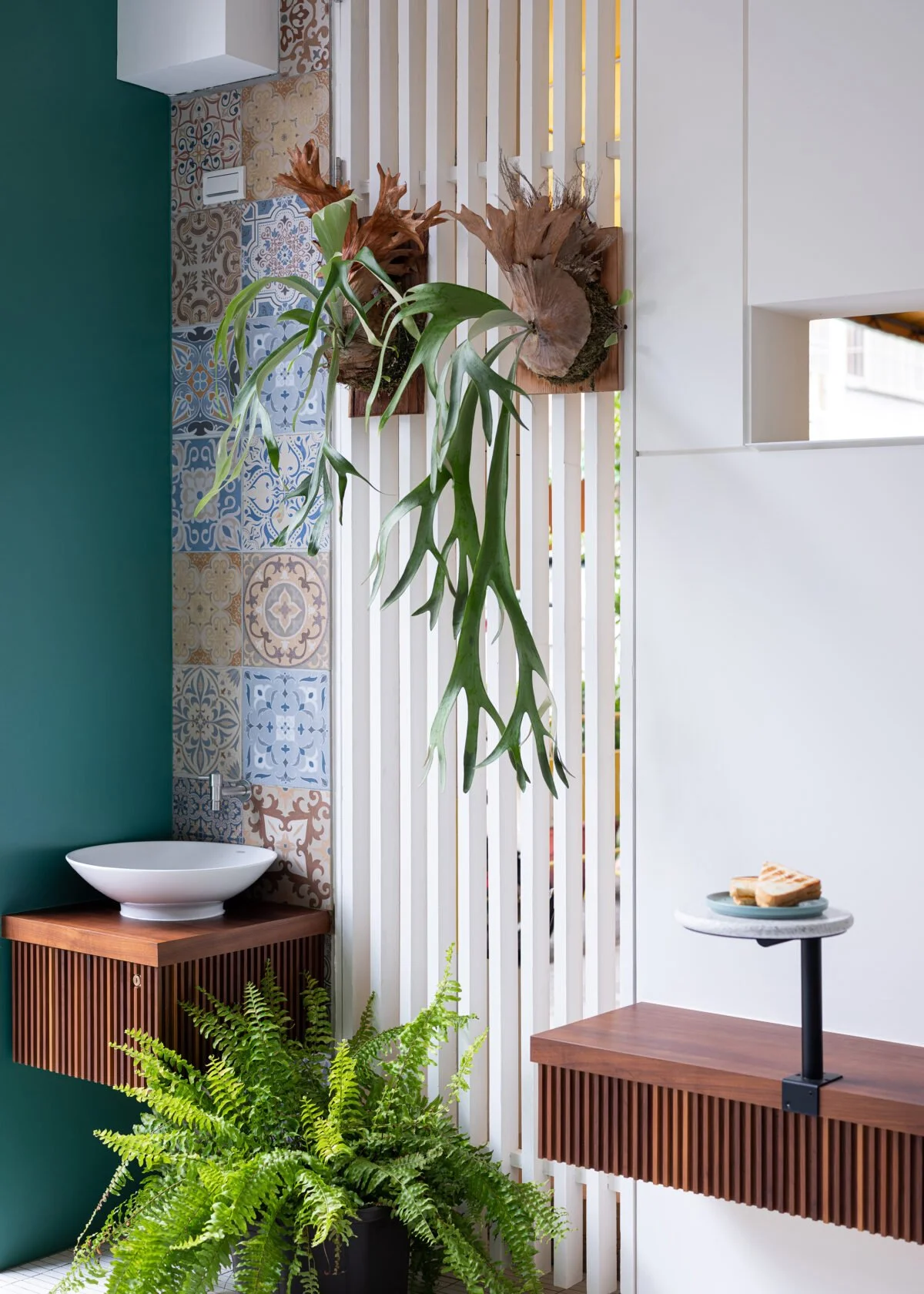 Interior of a modern space with a plant wall, decorative patterned tiles, a white vessel sink on a wooden slatted cabinet, and a small black-topped table with a plate of grilled sandwiches.