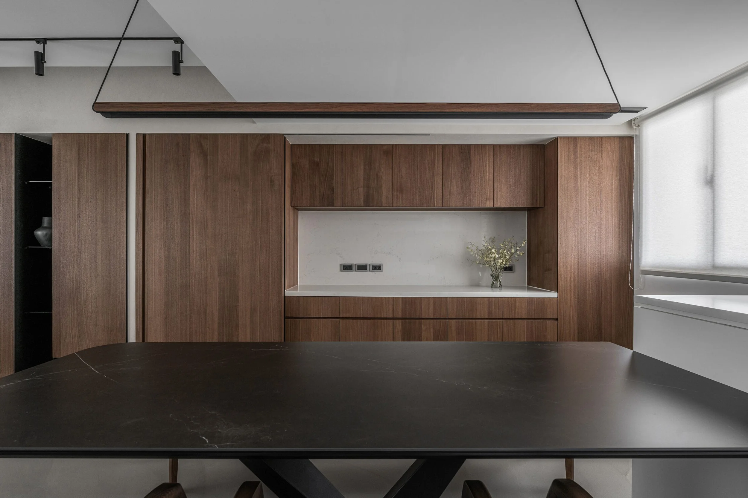 Modern interior with wooden cabinets, a corner shelf, and a vase of flowers on a white countertop. Part of a black table and a dark chair are visible in the foreground.